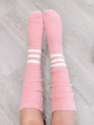 Pink fuzzy socks with white stripes, cozy and comfy, shown on legs against a wood floor.