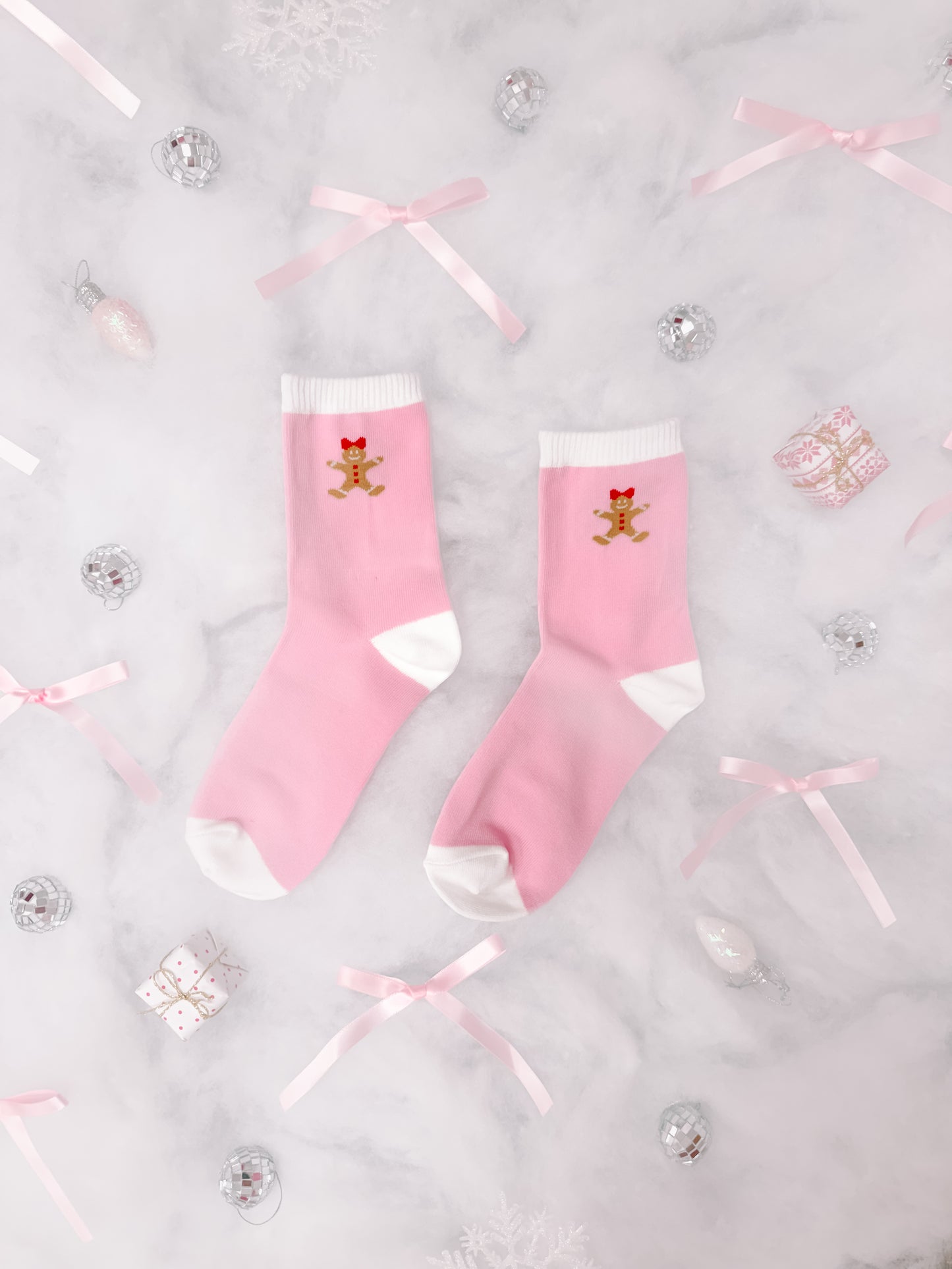 Pink crew socks with gingerbread man design, nestled amongst festive decorations on marble.