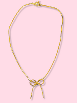 Gold chain necklace with a delicate bow pendant against a pink background.