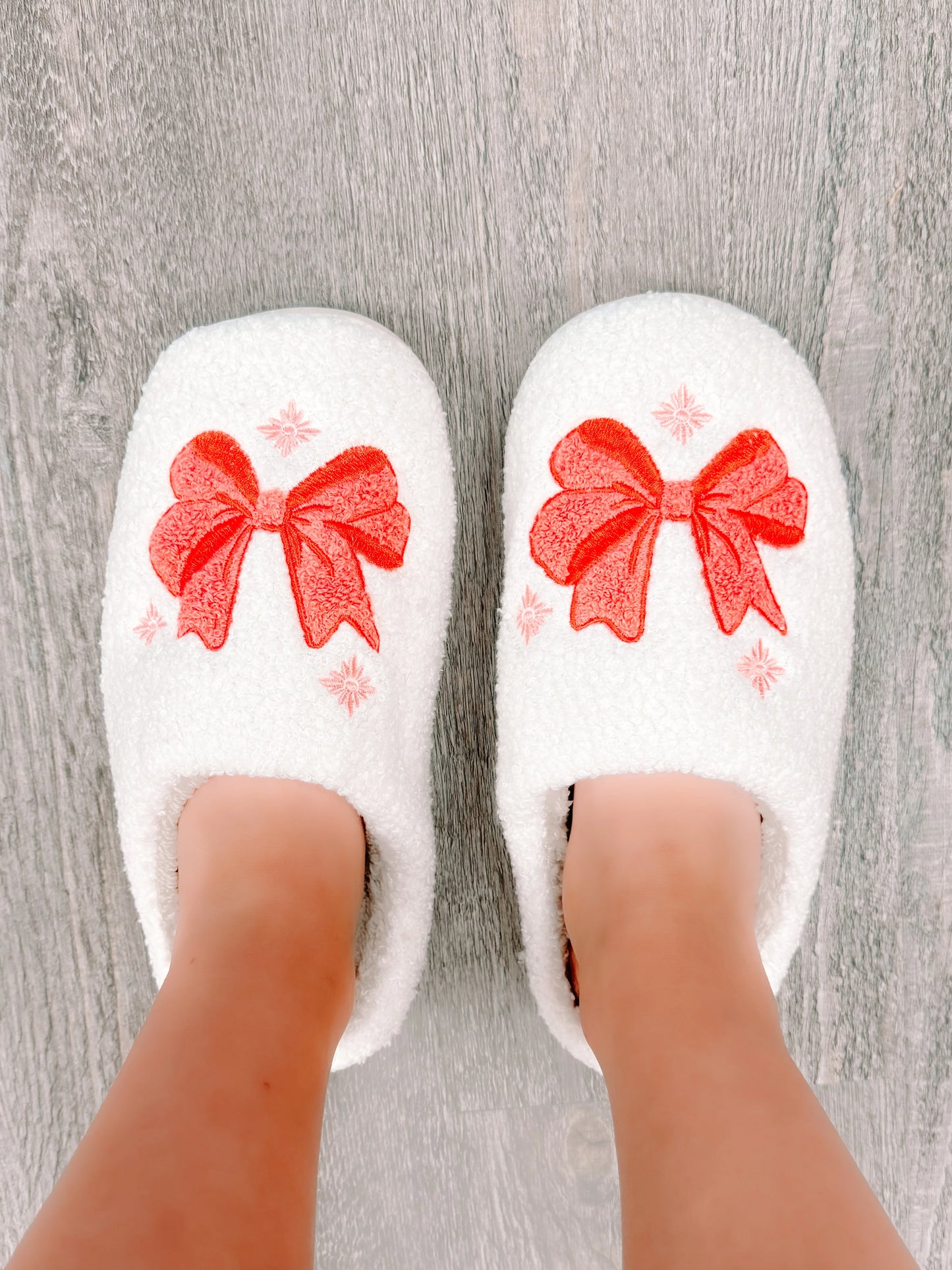 White fuzzy slippers with coral bows, worn on a wood floor.