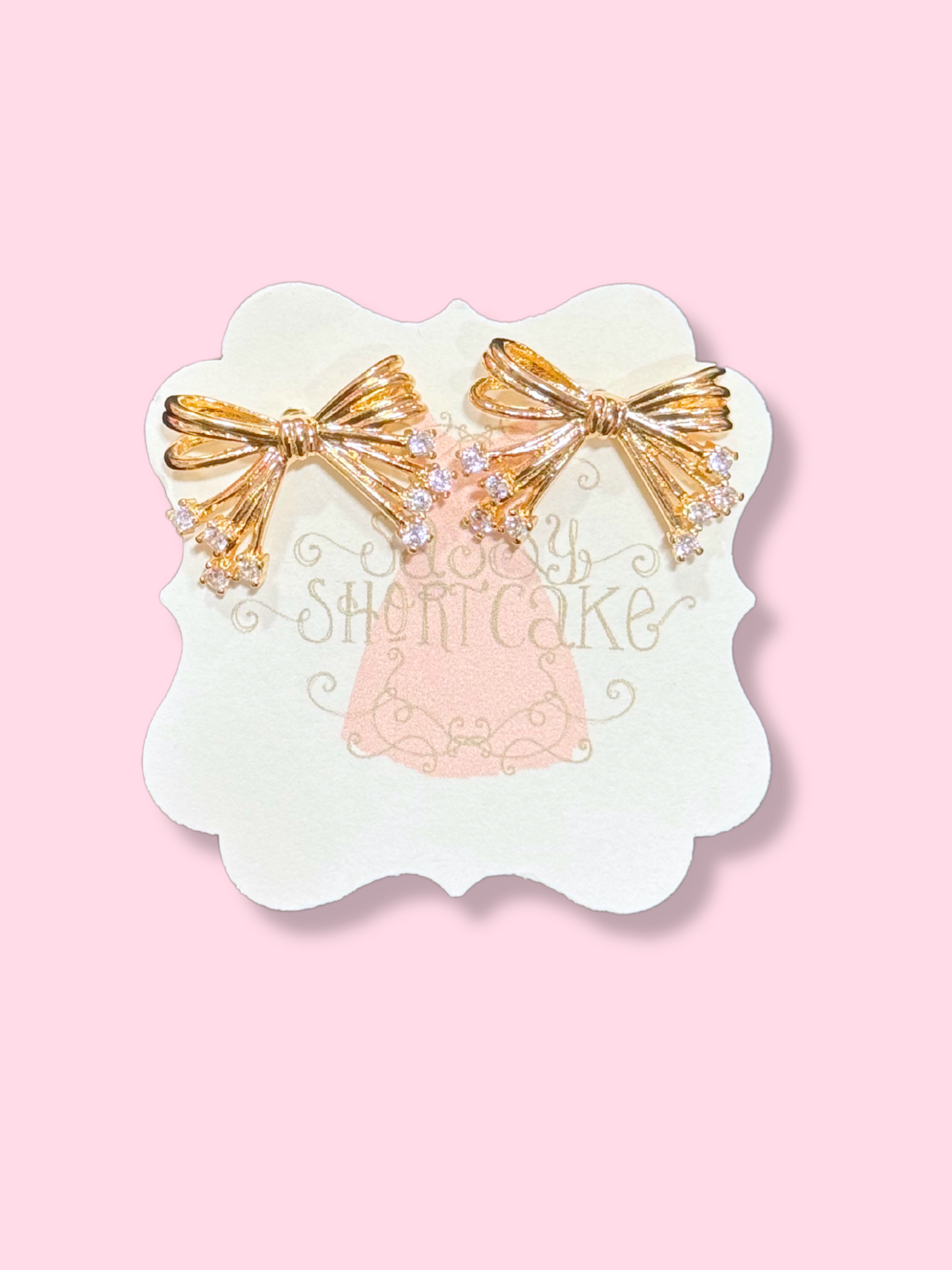 Gold Lucille Bow earrings with sparkling crystals, displayed on a pink background.
