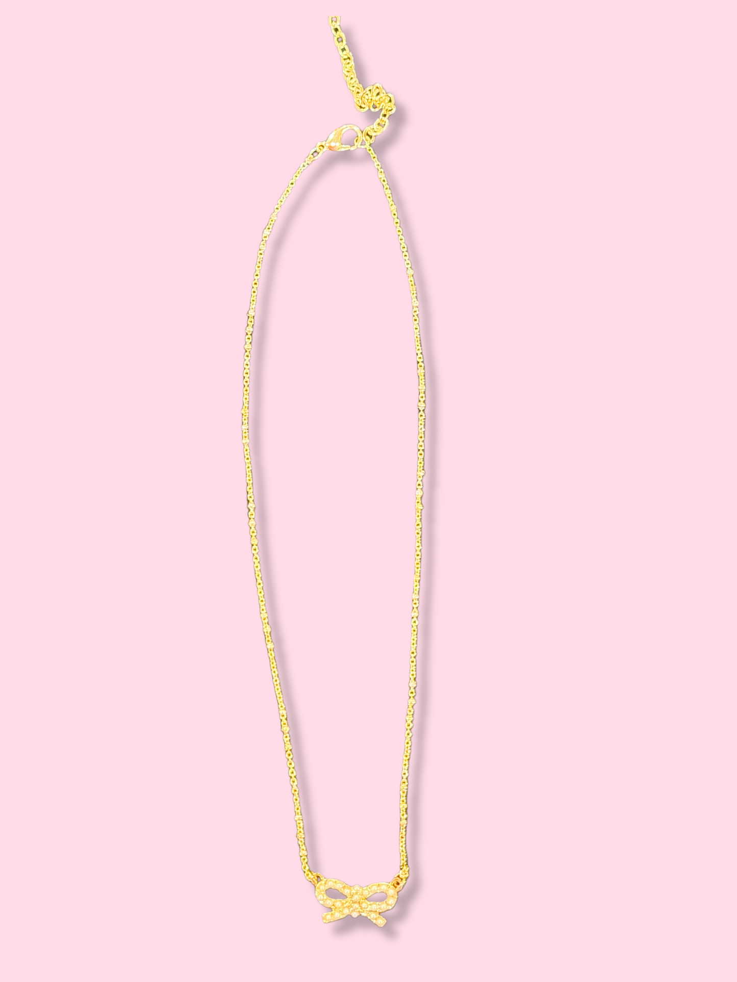 Gold chain necklace with a small pearl bow pendant against a pink background.