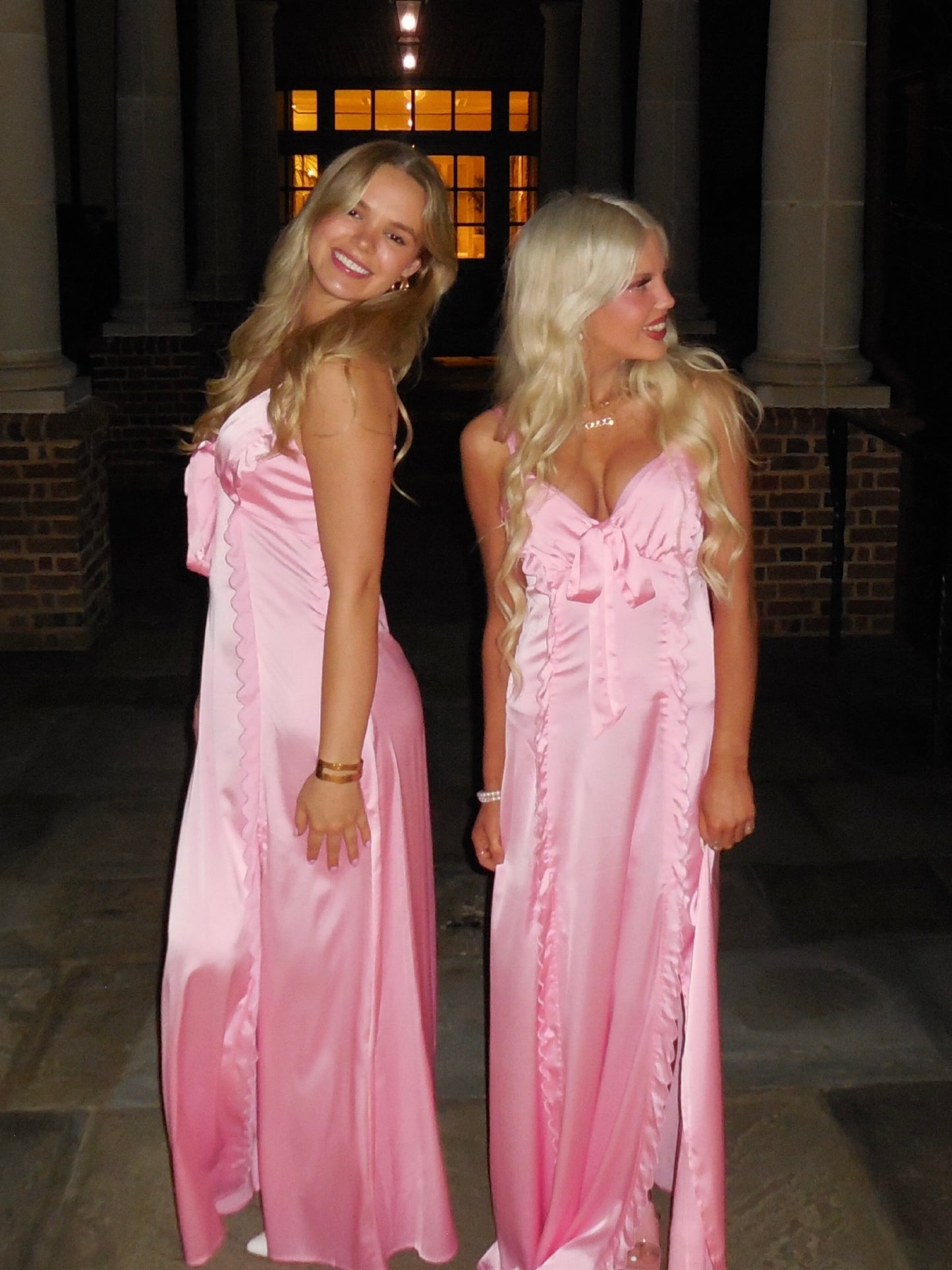 Two women in matching pink satin maxi dresses, posing outdoors at night.