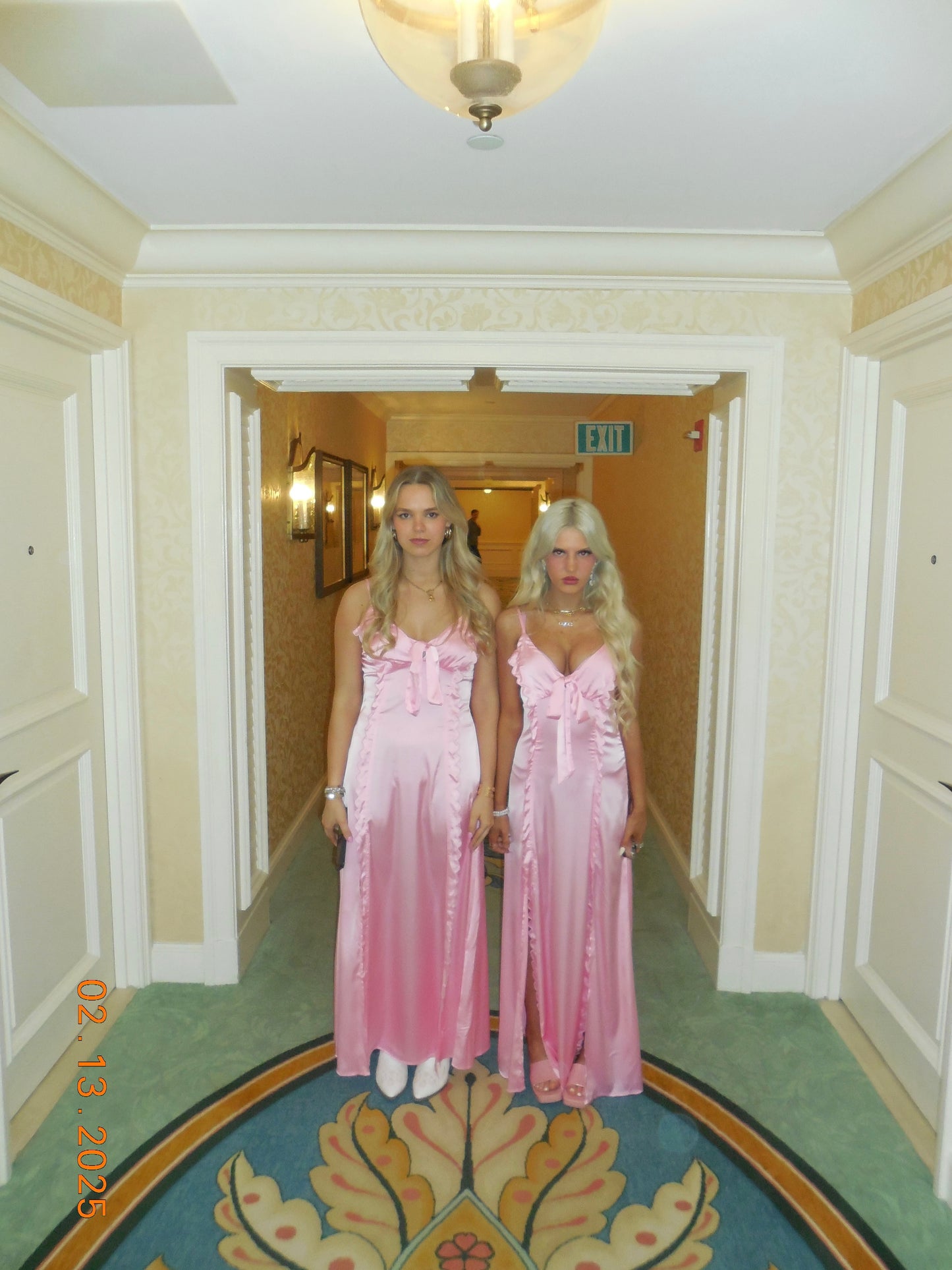 Two women in pink satin gowns pose in a hotel hallway.