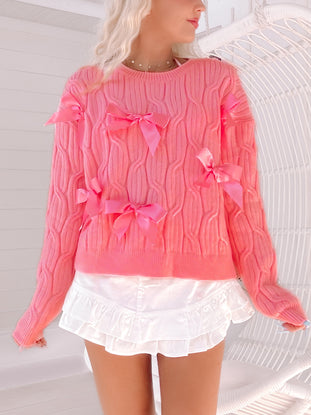 Pink cable-knit sweater adorned with pink satin bows, modeled against a white background.