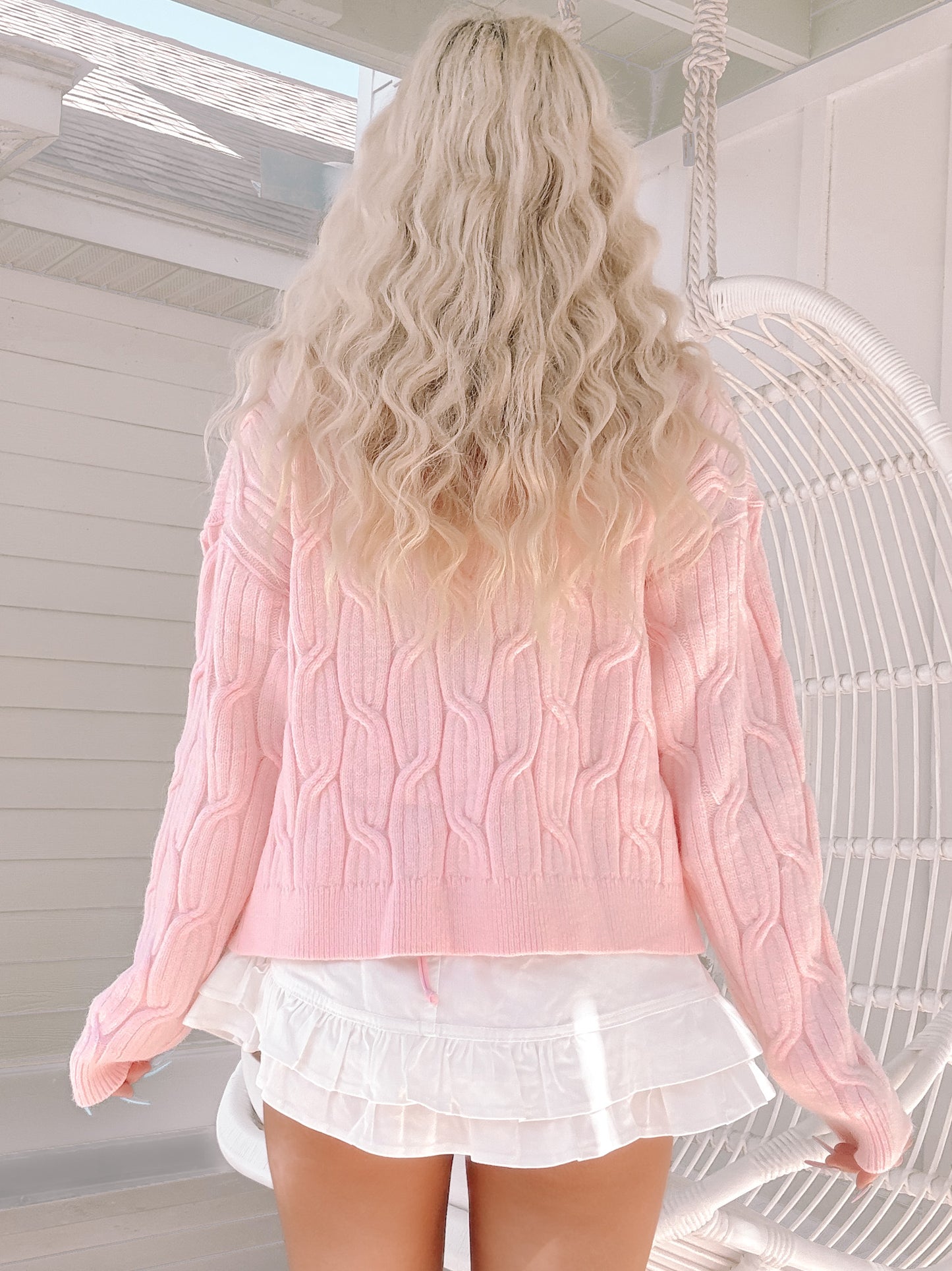 Light pink cable-knit sweater, shown from behind on a model near a white wicker chair.