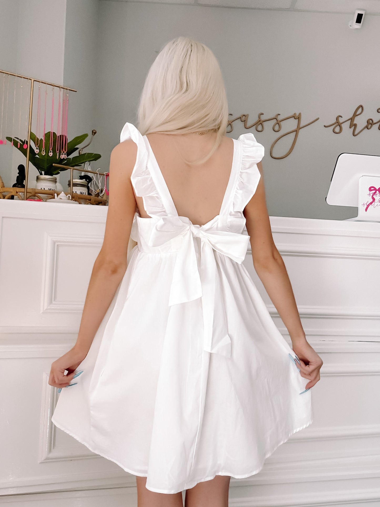 White eyelet ruffle mini dress with bow back detail, shown on model in boutique.