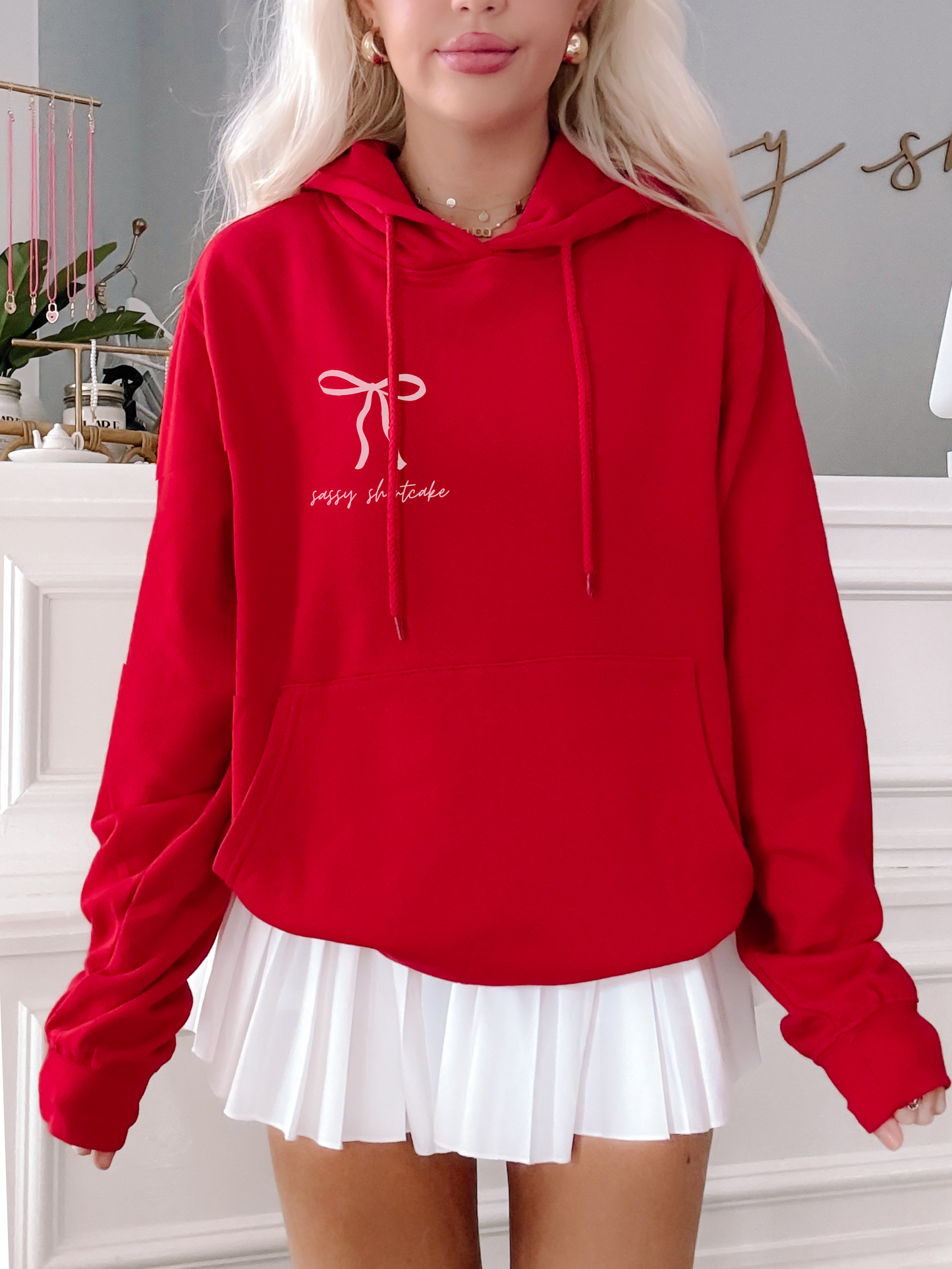 Red oversized hoodie with a pink bow and text graphic, modeled over a white pleated skirt.
