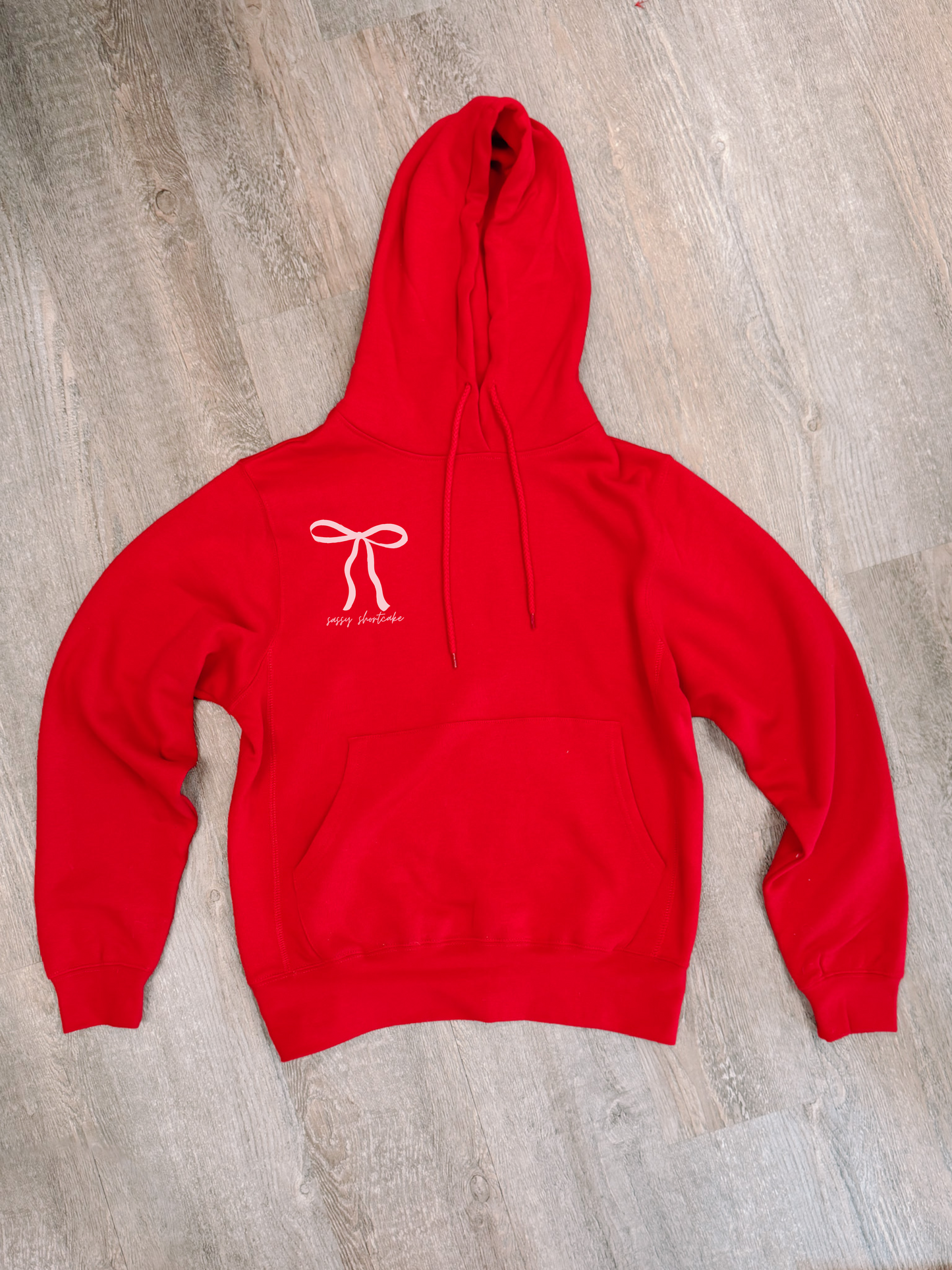 Red hoodie featuring a bow and "sassy shortcake" text, displayed on a wood floor.