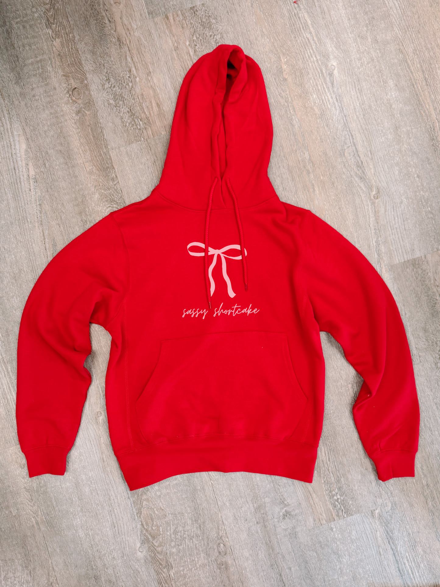 Red hoodie with a bow and “Sassy Shortcake” design, laid flat on wood.