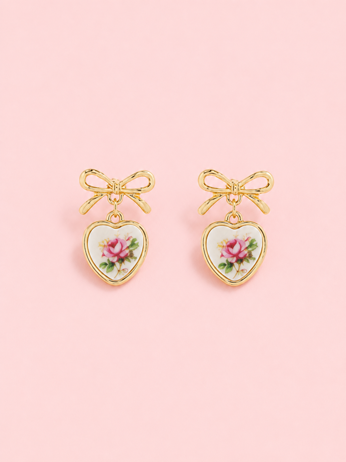 Sweet Bloom Pink Bow Earrings from Sassy Shortcake: Gold bow earrings with ceramic heart charms featuring a pink rose print on a light pink background.
