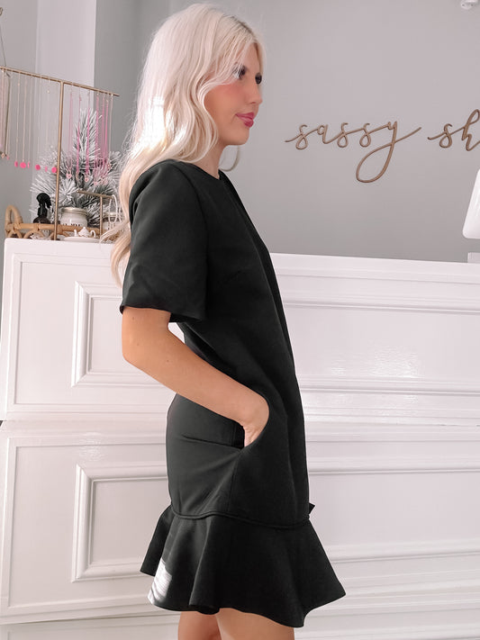 Black short-sleeve dress with ruffled hem, shown on model against white wall.