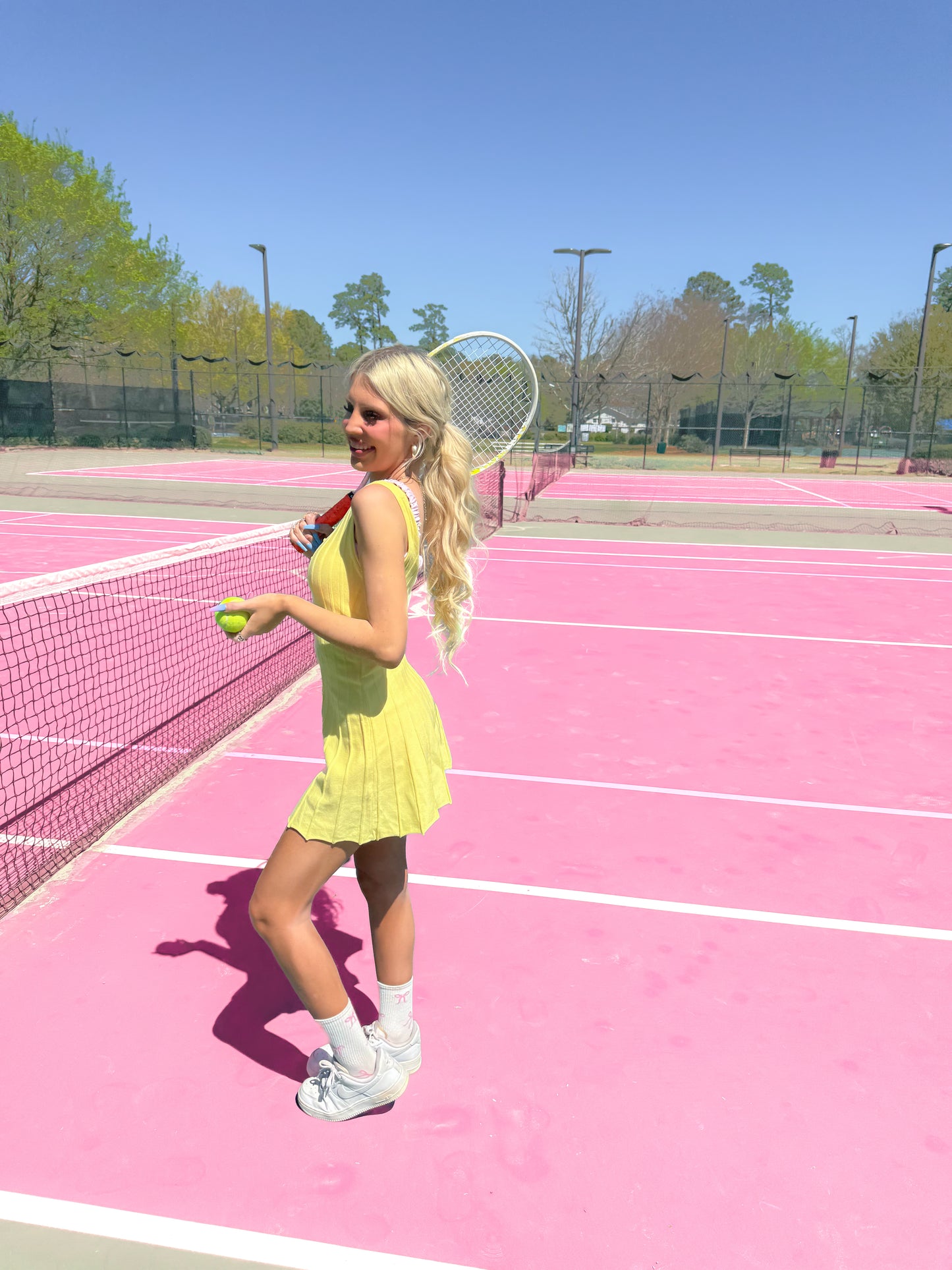 Woman in yellow tennis dress on pink court, holding tennis racket and ball.