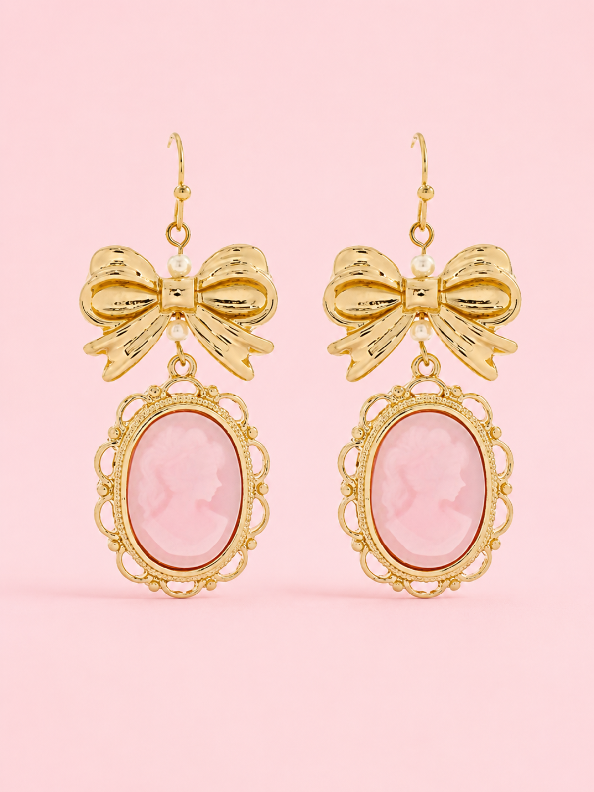 Vintage Muse Bow Earrings from Sassy Shortcake featuring gold-tone bow details, pink oval cameo charms, and fish hook backs on a light pink background.