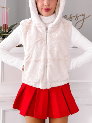 Cream faux fur zip-up vest with attached hoodie, styled with a red pleated mini skirt.