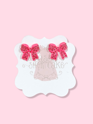 Pink bow stud earrings adorned with sparkling crystals, displayed on a pink background.