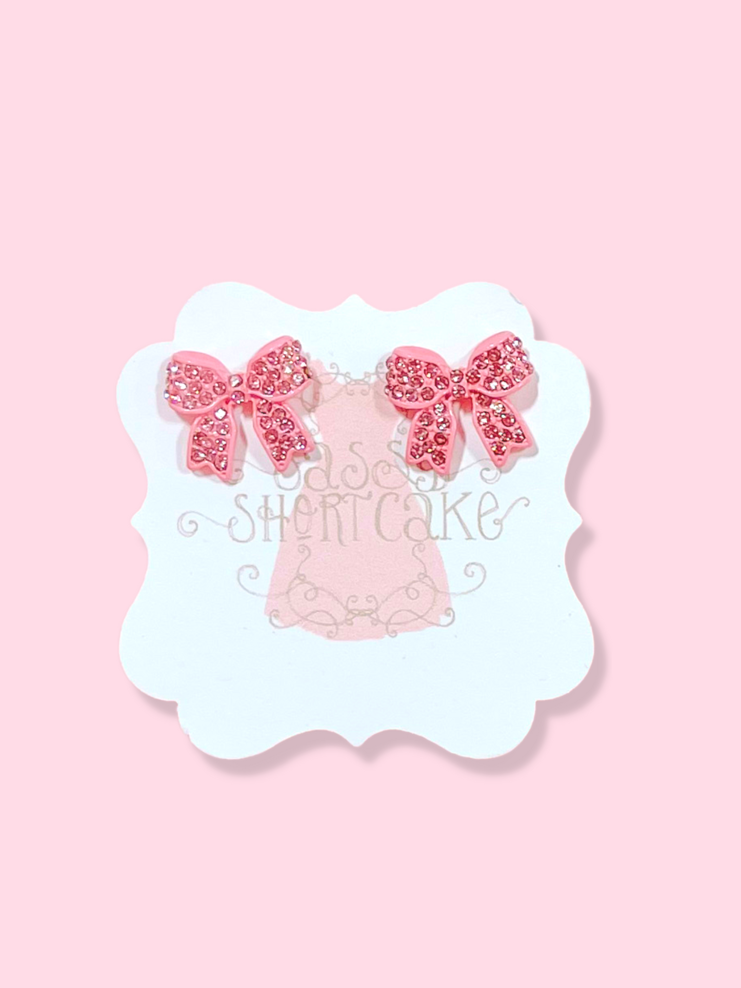 Pink bow stud earrings adorned with sparkling crystals, displayed on a light pink background.