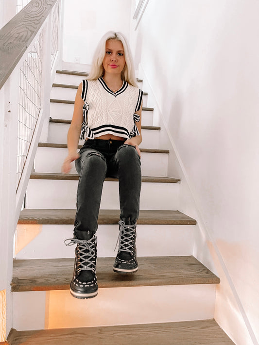 Cream cable knit vest styled with black jeans, worn indoors on a staircase.