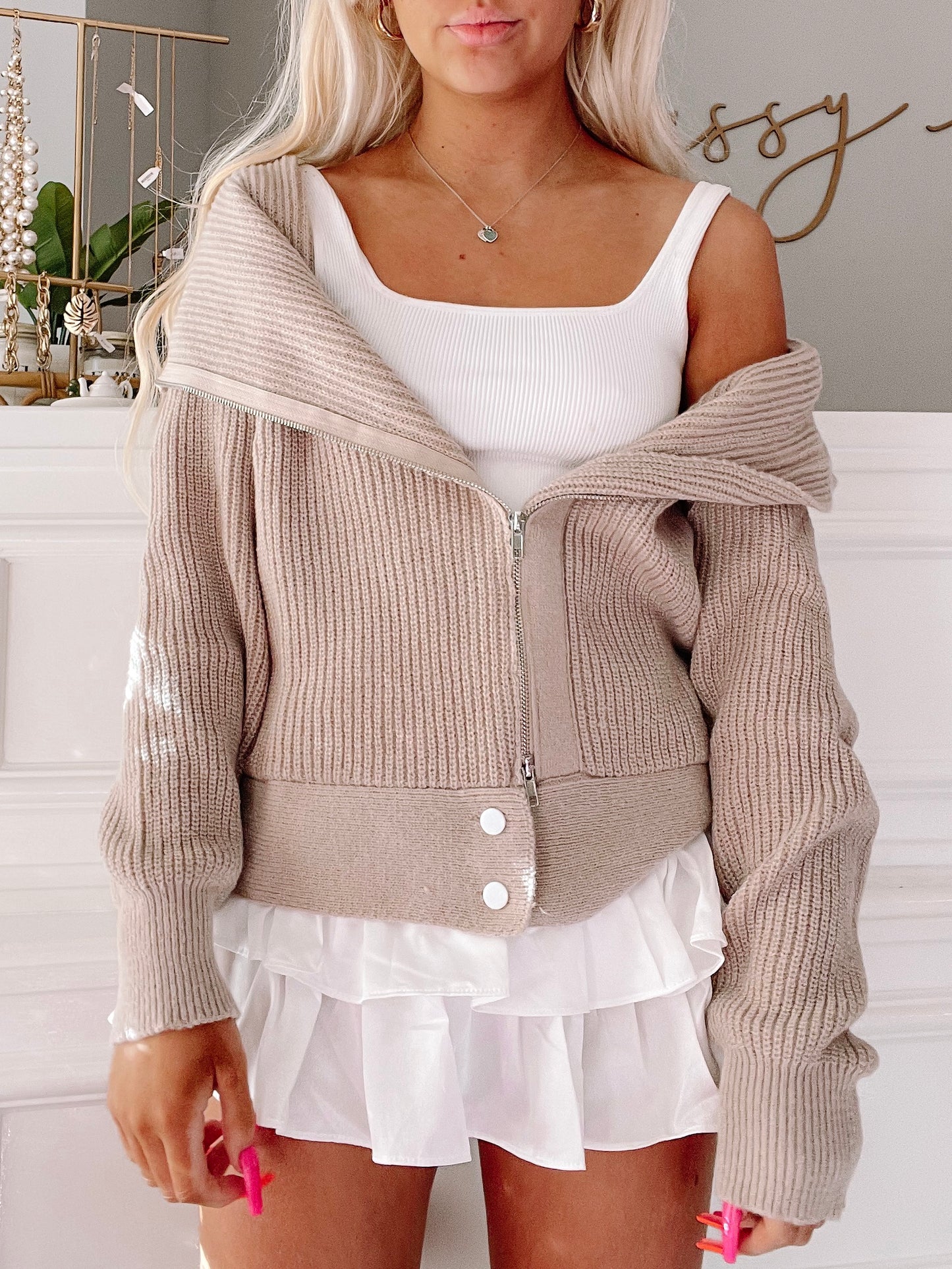 Beige zip-up knit cardigan with ribbed texture, worn over a white tank top and layered skirt.