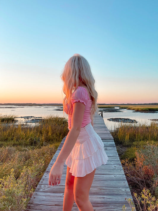 Woman in pink top and white tiered skirt on a boardwalk at sunset over marshland.