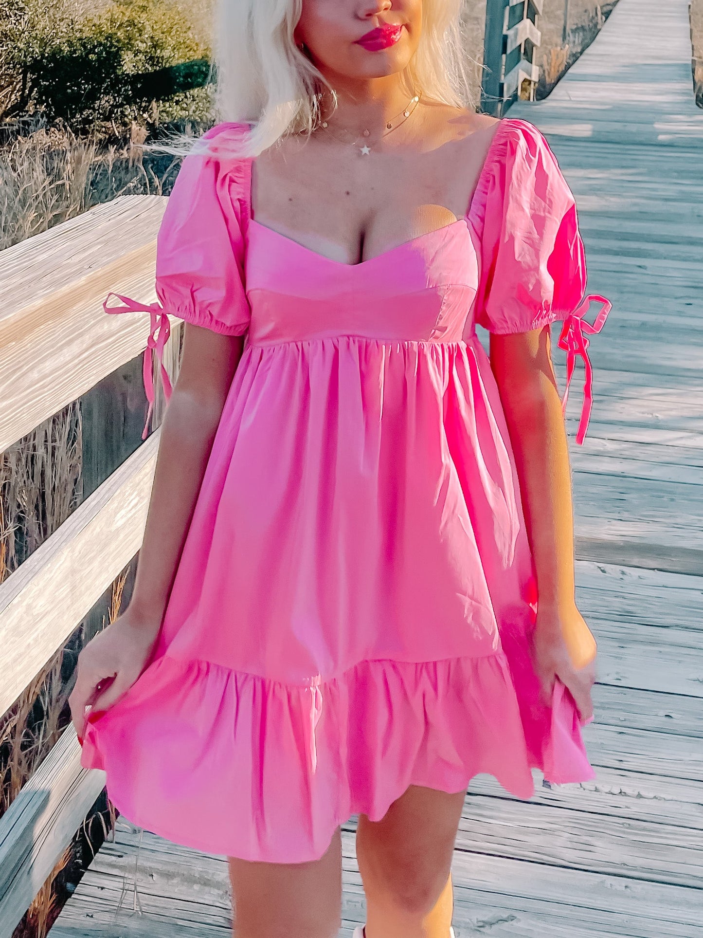 Hot pink mini dress with puff sleeves and tie detailing, worn outdoors on a boardwalk.
