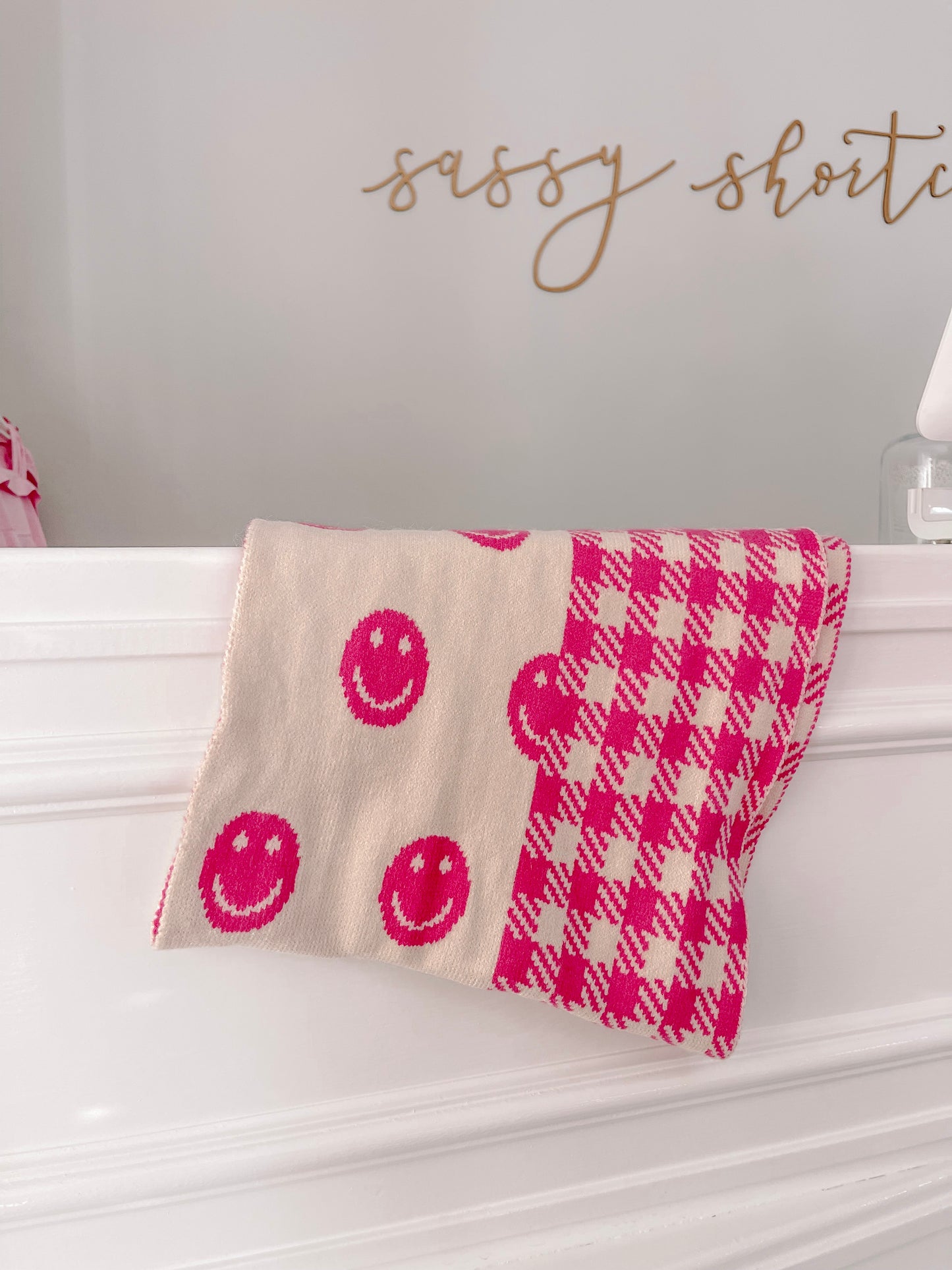 Cream and hot pink knitted scarf with smiley faces and houndstooth pattern, draped on white mantel.