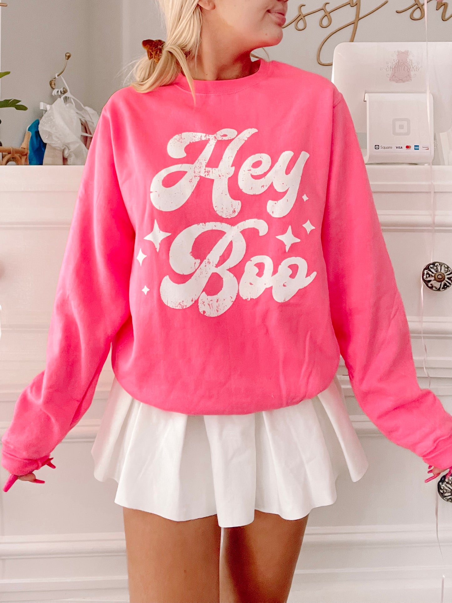 Hot pink crewneck sweatshirt with a retro "Hey Boo" graphic and sparkling details, modeled with a white pleated mini skirt.