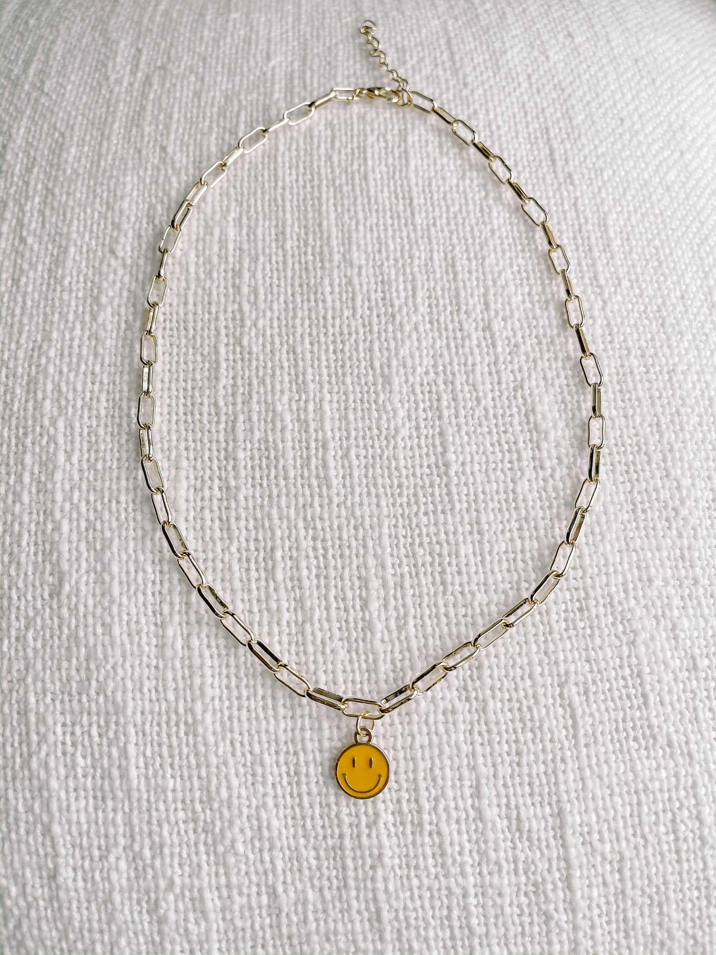 Gold chain necklace with cheerful yellow smiley face charm, displayed on white textured fabric.