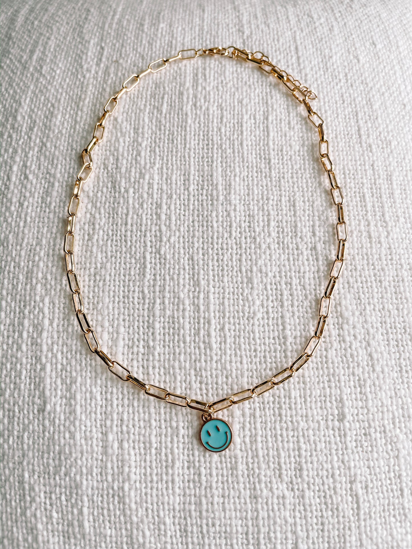 Gold chain necklace with light blue smiley face charm, displayed on textured white fabric.