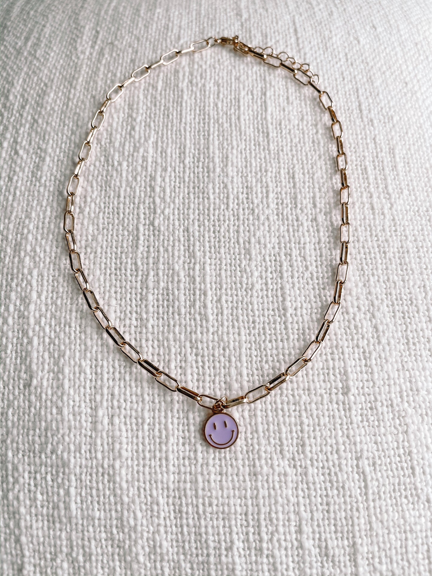 Gold chain necklace with lavender smiley face charm on textured white fabric