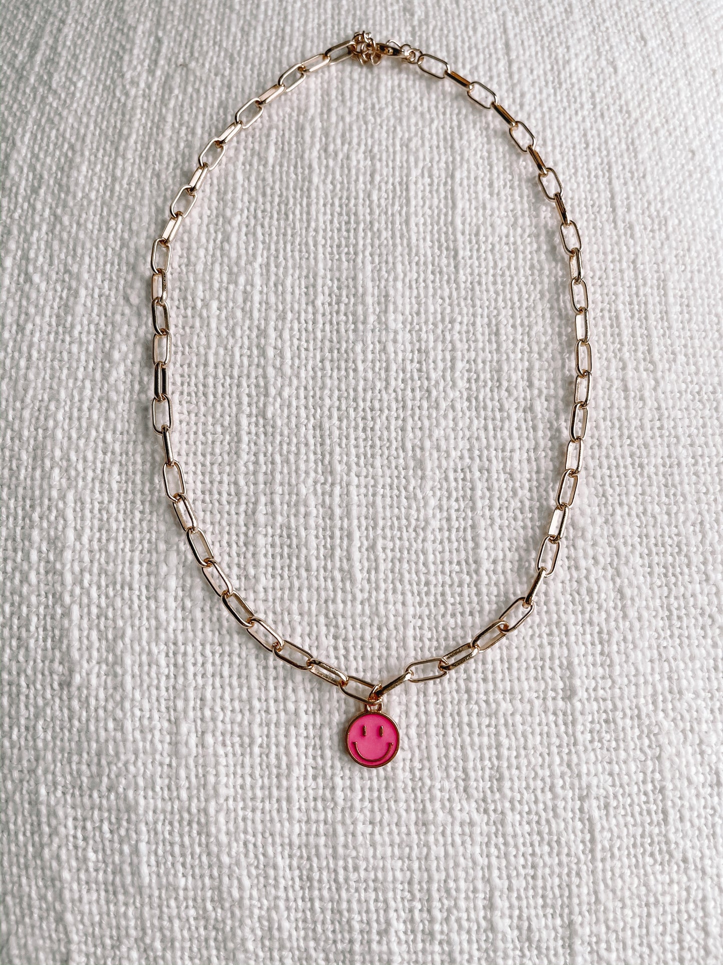 Gold chain necklace with a pink smiley face charm, displayed on a textured white surface.