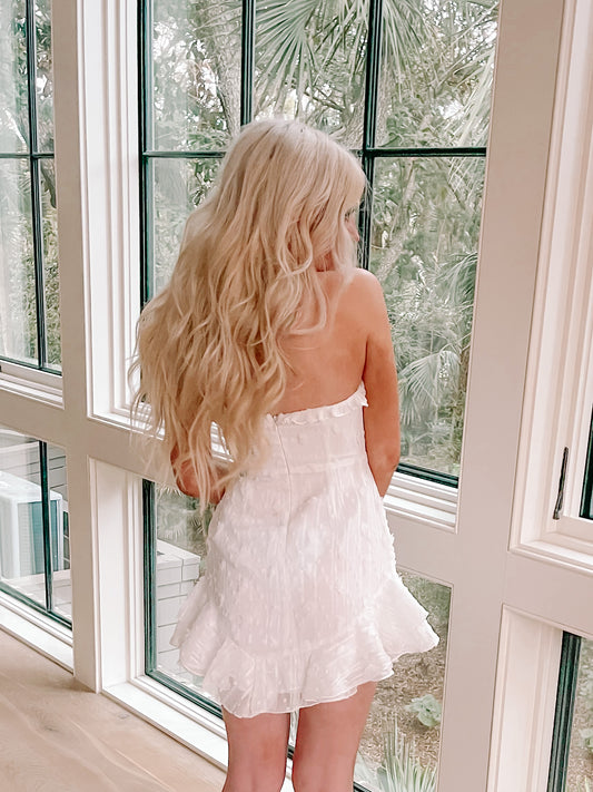 White eyelet mini dress with ruffled hem, shown from behind near a window.