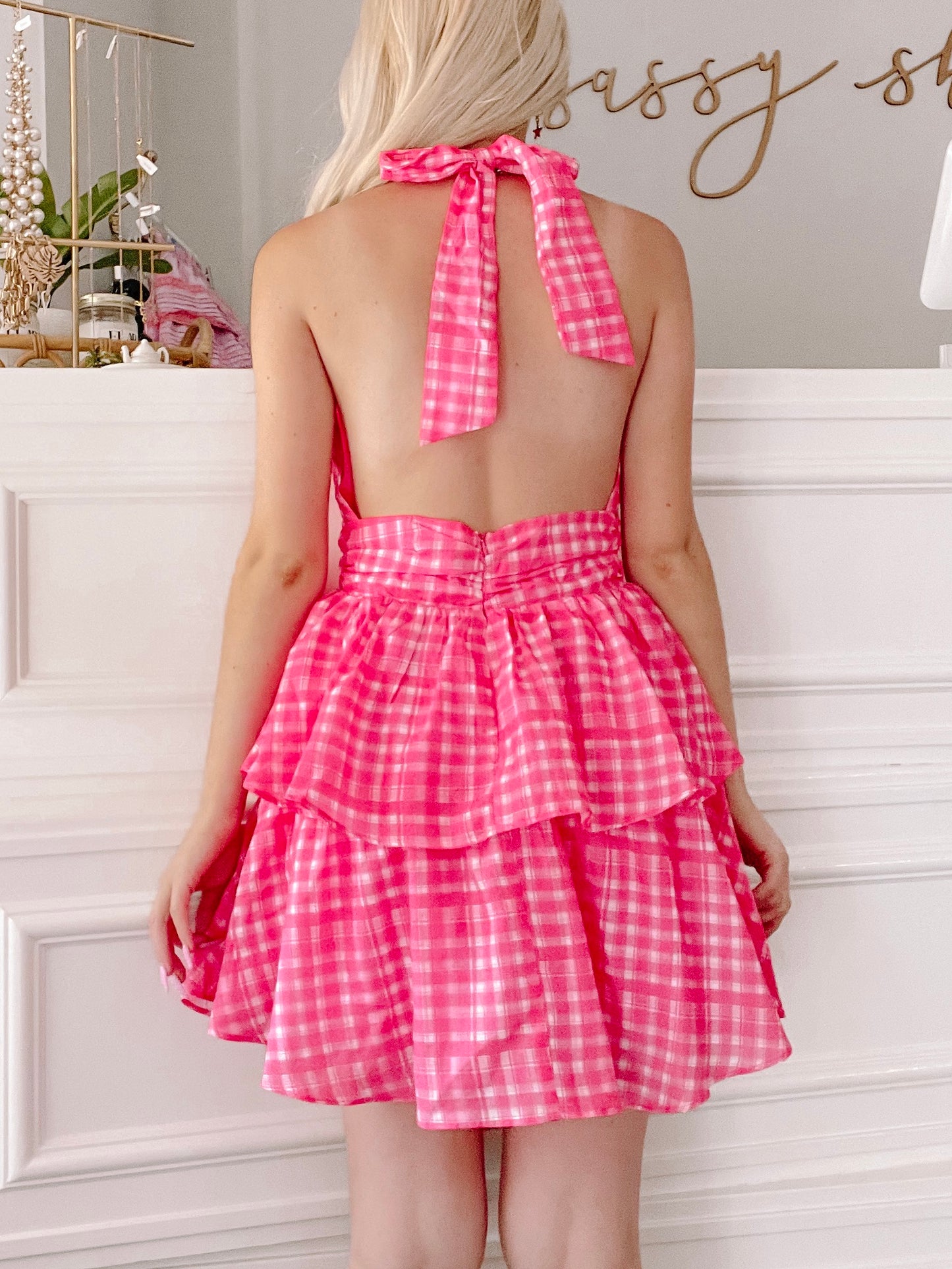Pink gingham halter mini dress with tiered ruffle skirt, shown from the back against a white wall.