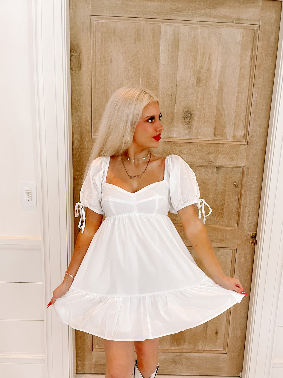 White puff-sleeve mini dress with tiered skirt, shown on model against light wood door.