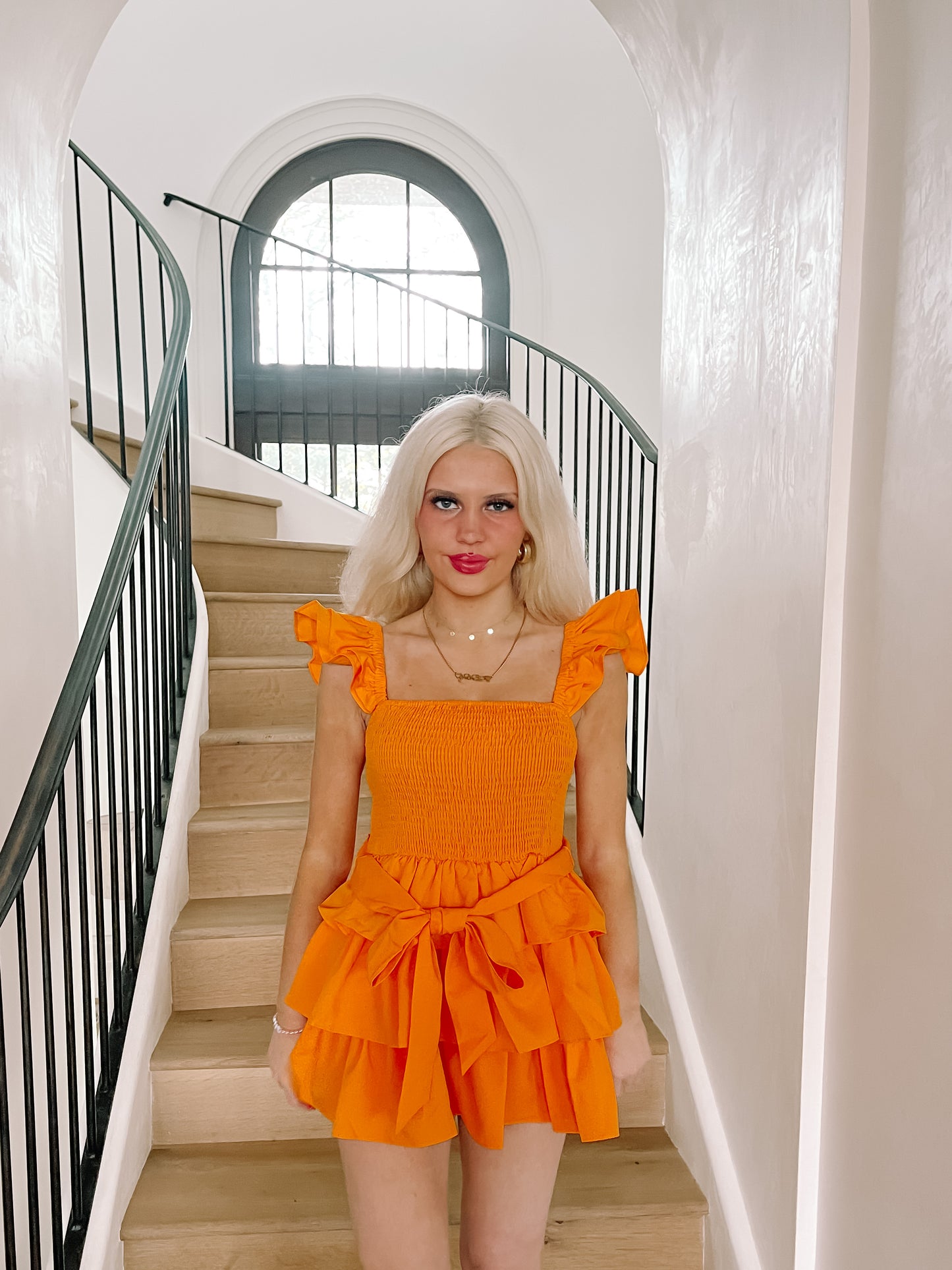 Bright orange tiered mini dress with ruffle sleeves, modeled on a staircase in a stylish home.