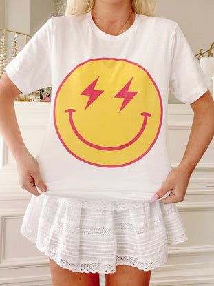 White cotton tee showcasing a yellow smiley face graphic with pink lightning bolt eyes; model shown wearing it with a white eyelet skirt.