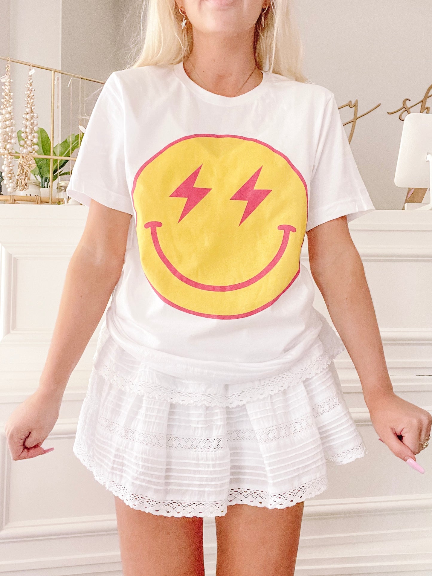 White cotton t-shirt featuring a yellow smiley face graphic with pink lightning bolts; model also wears a white eyelet skirt.