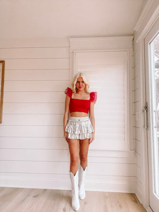 Woman in red crop top and silver skirt with white cowboy boots, posing against white shiplap wall.