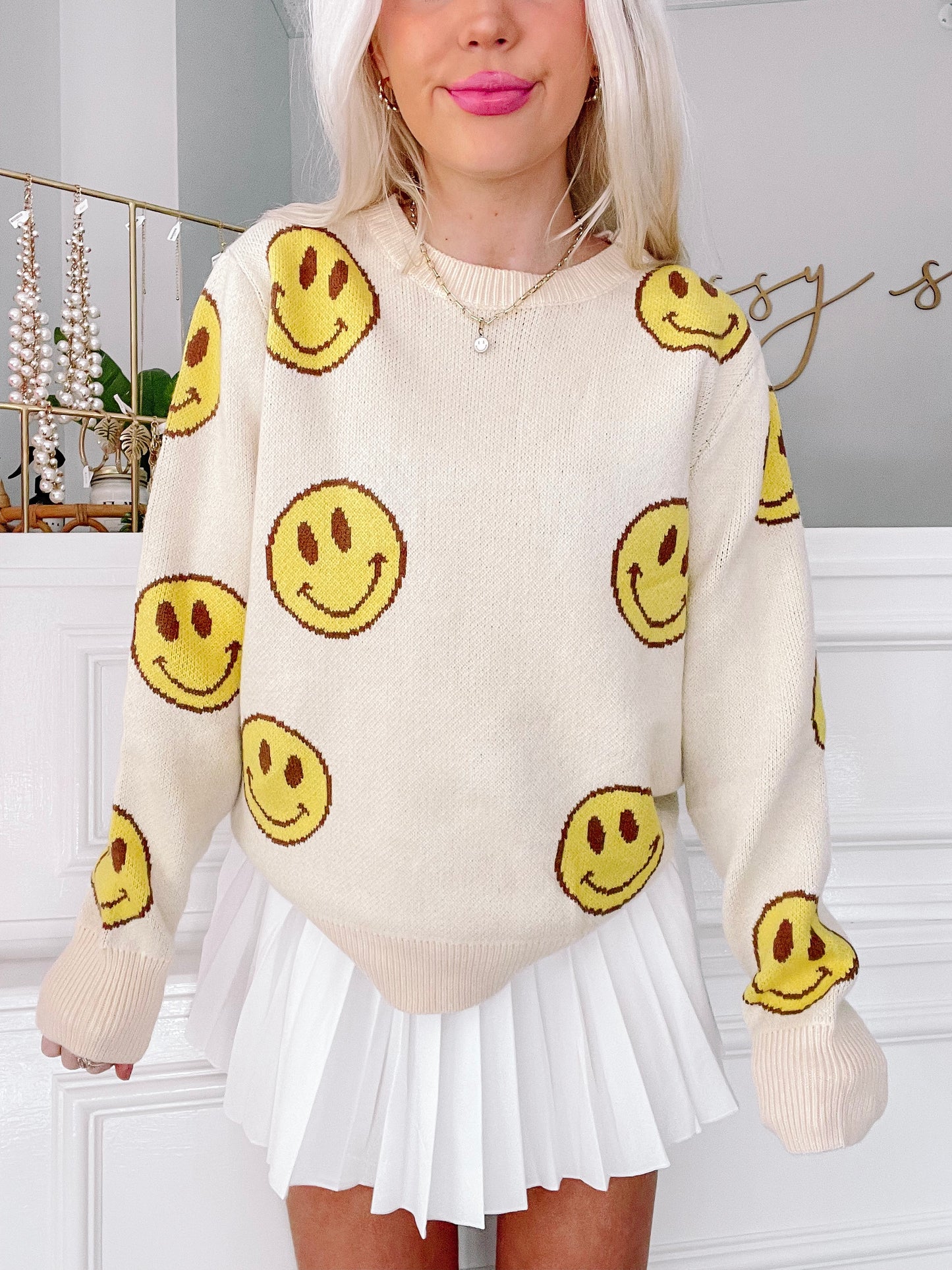 Cream-colored knit sweater adorned with yellow smiley faces, modeled with a white pleated skirt.