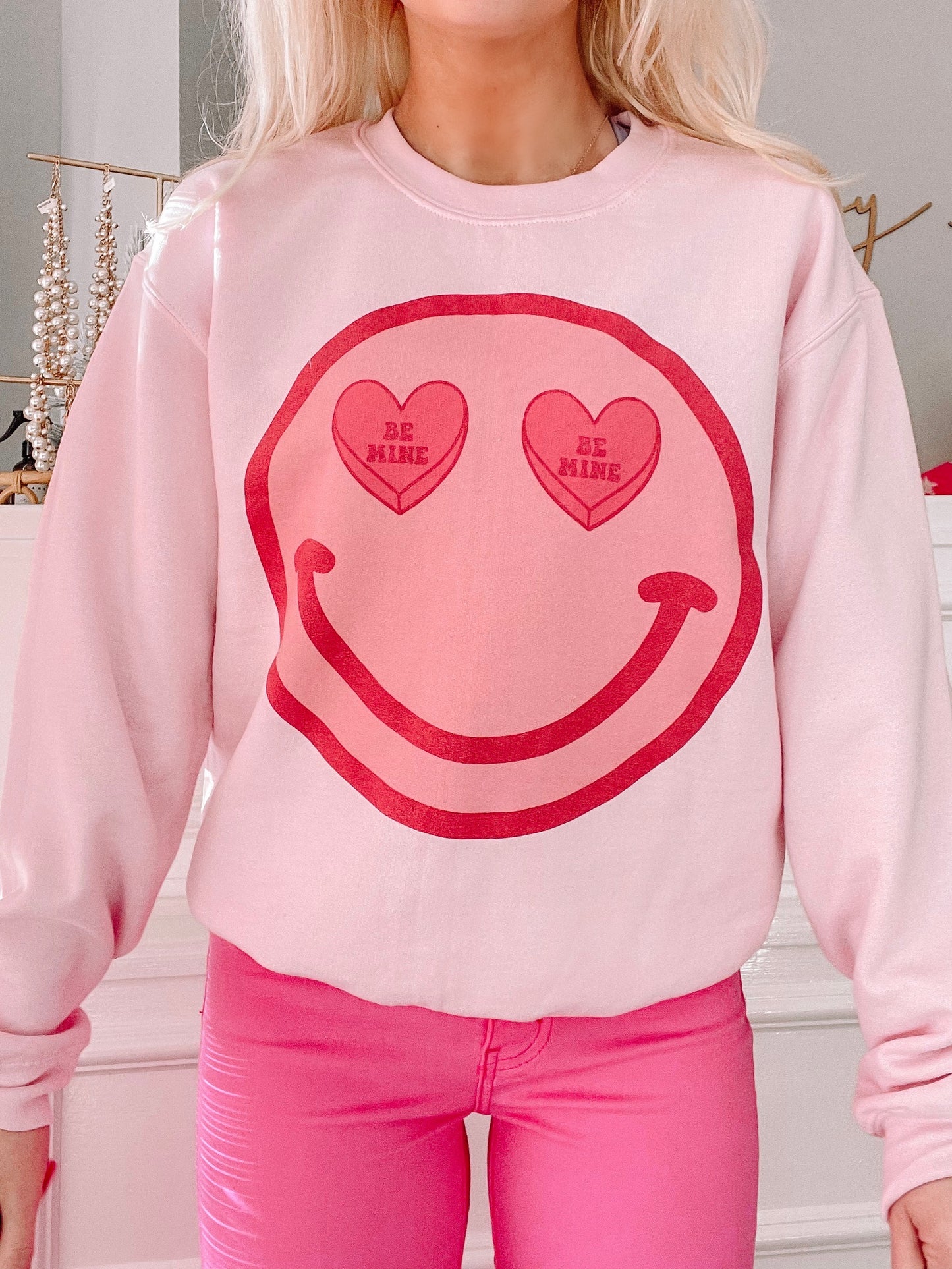 Pink crewneck sweatshirt featuring a smiling face graphic with "Be Mine" candy hearts for eyes.