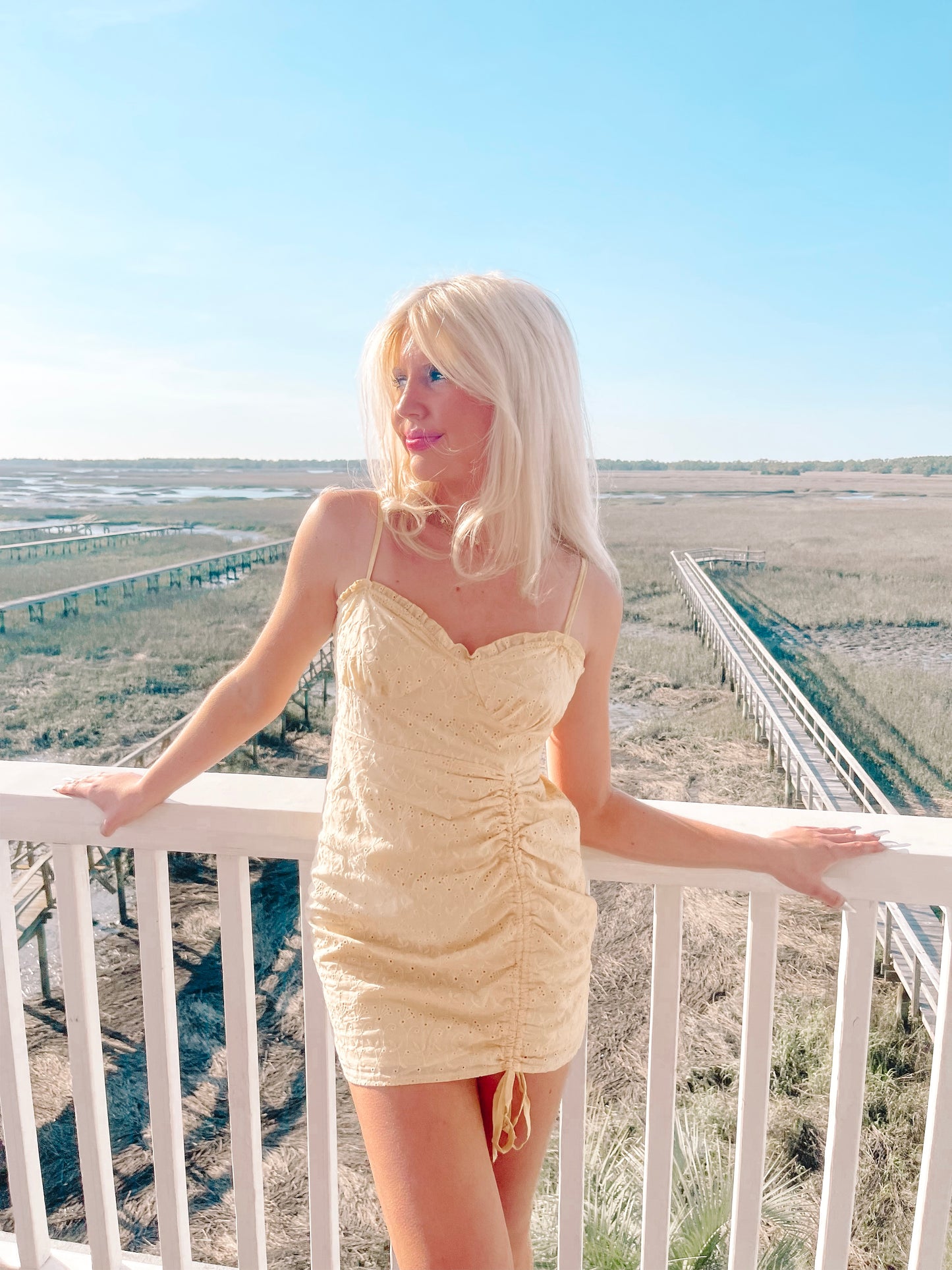 Pale yellow eyelet mini dress, cinched waist, worn outdoors on a sunny day overlooking marshland.