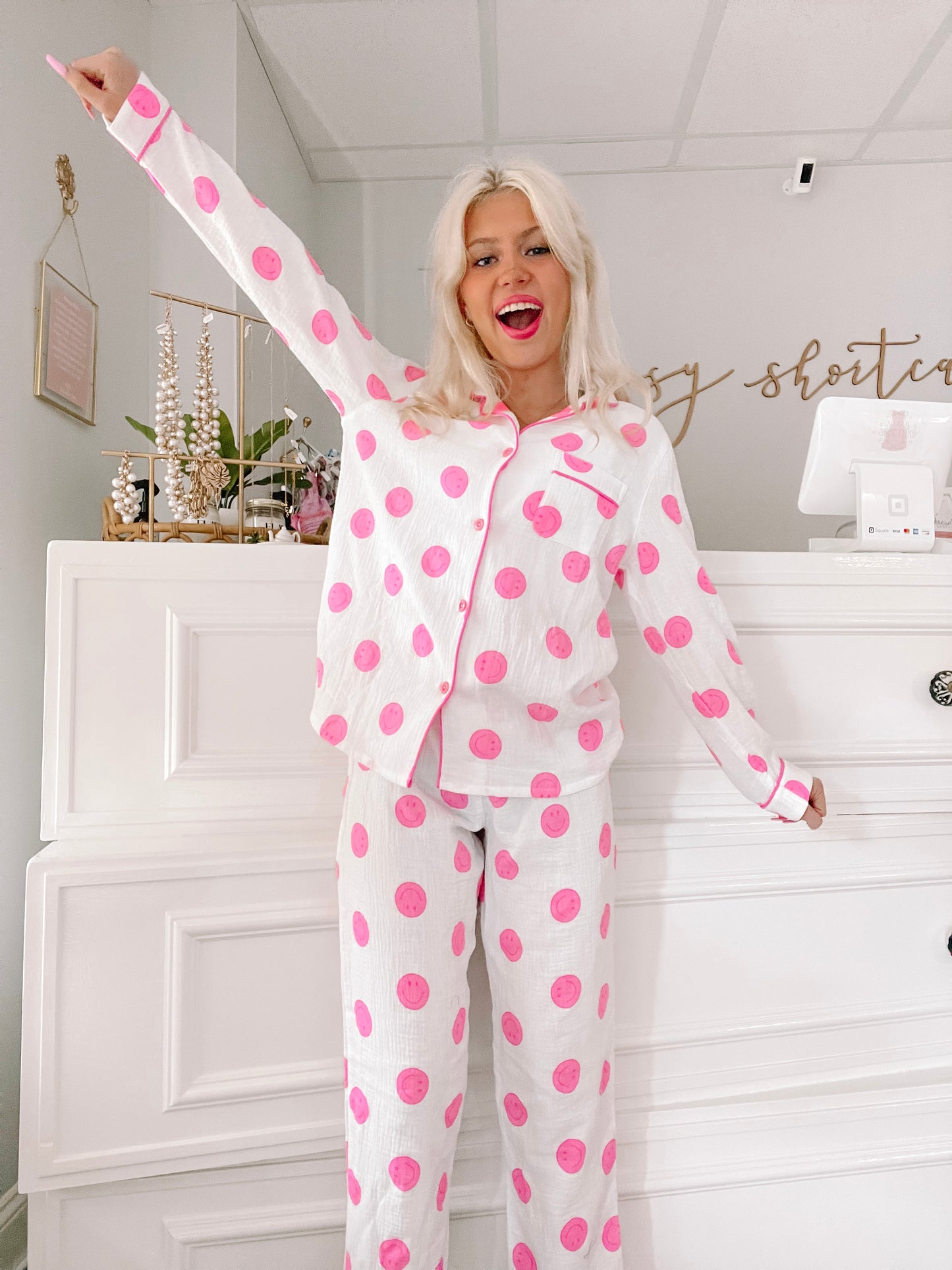 White pajama set with pink smiley faces, long sleeves, and pants; model shown in a bright room.