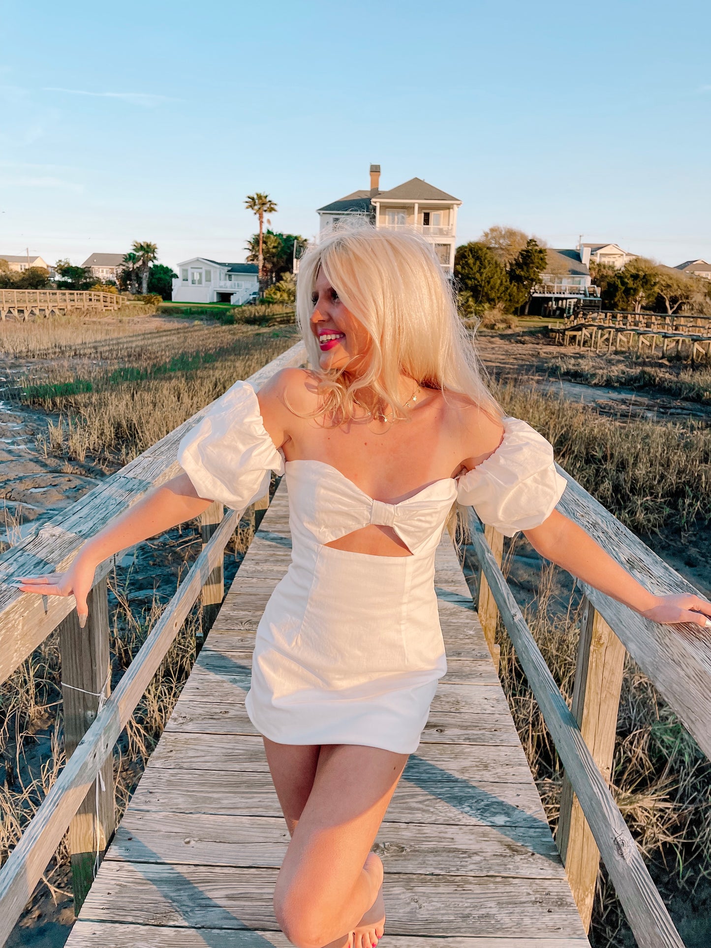 White off-the-shoulder mini dress with a bow detail, modeled outdoors on a wooden boardwalk.