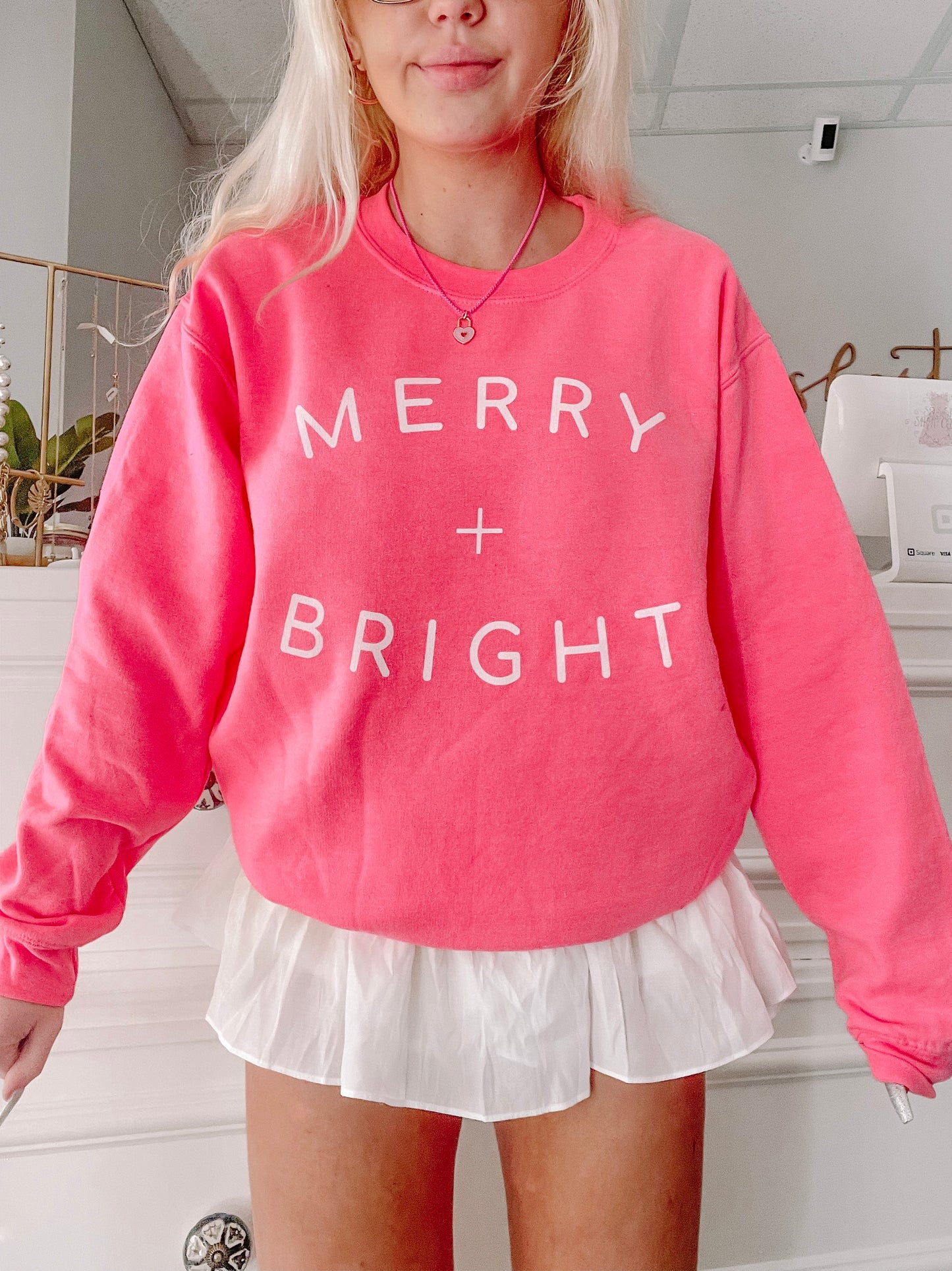 Pink crewneck sweatshirt with "Merry + Bright" in white lettering, modeled with a white skirt.