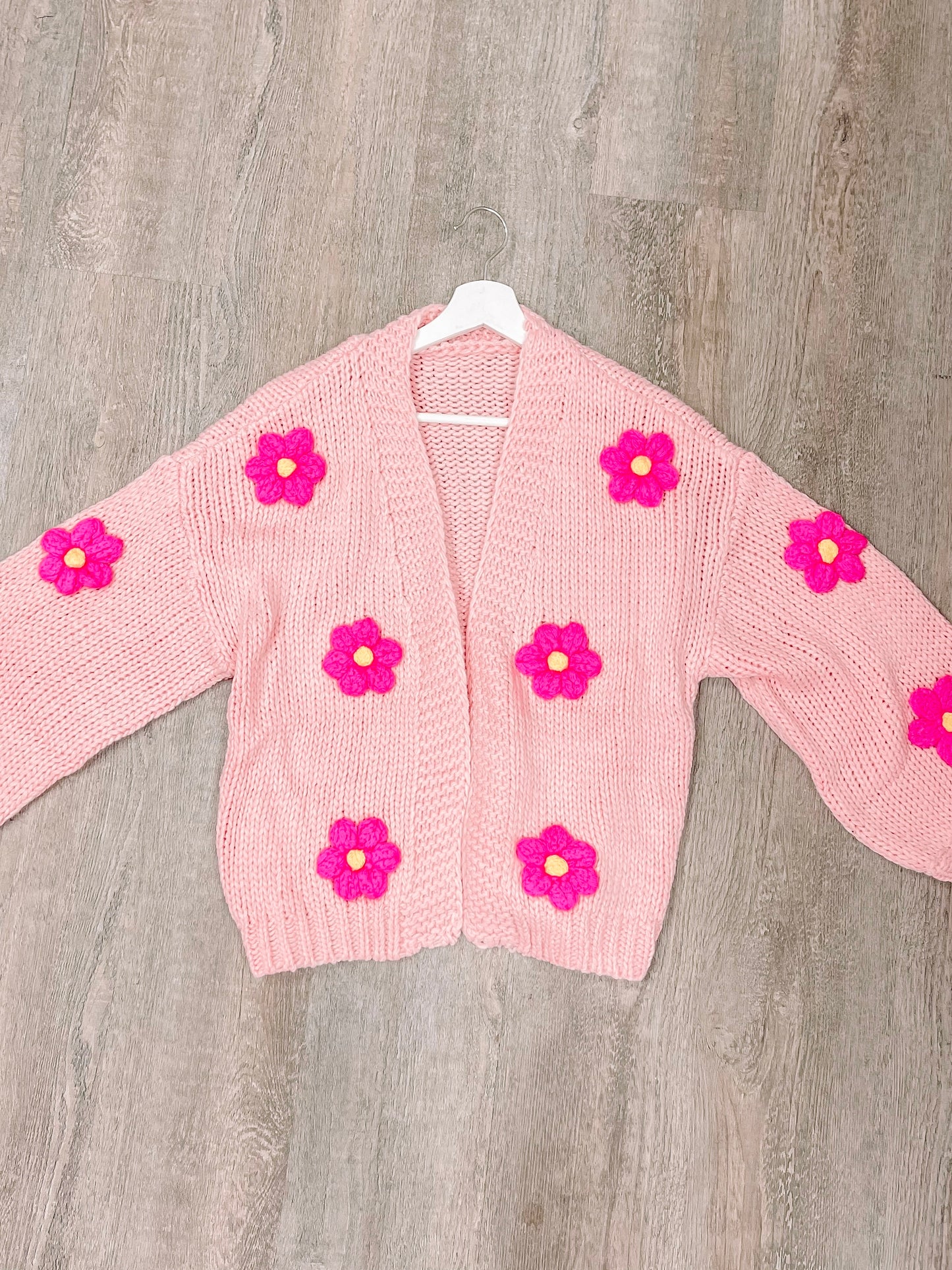 Pink knit cardigan adorned with bright pink flowers, hanging on a white hanger against a wood background.