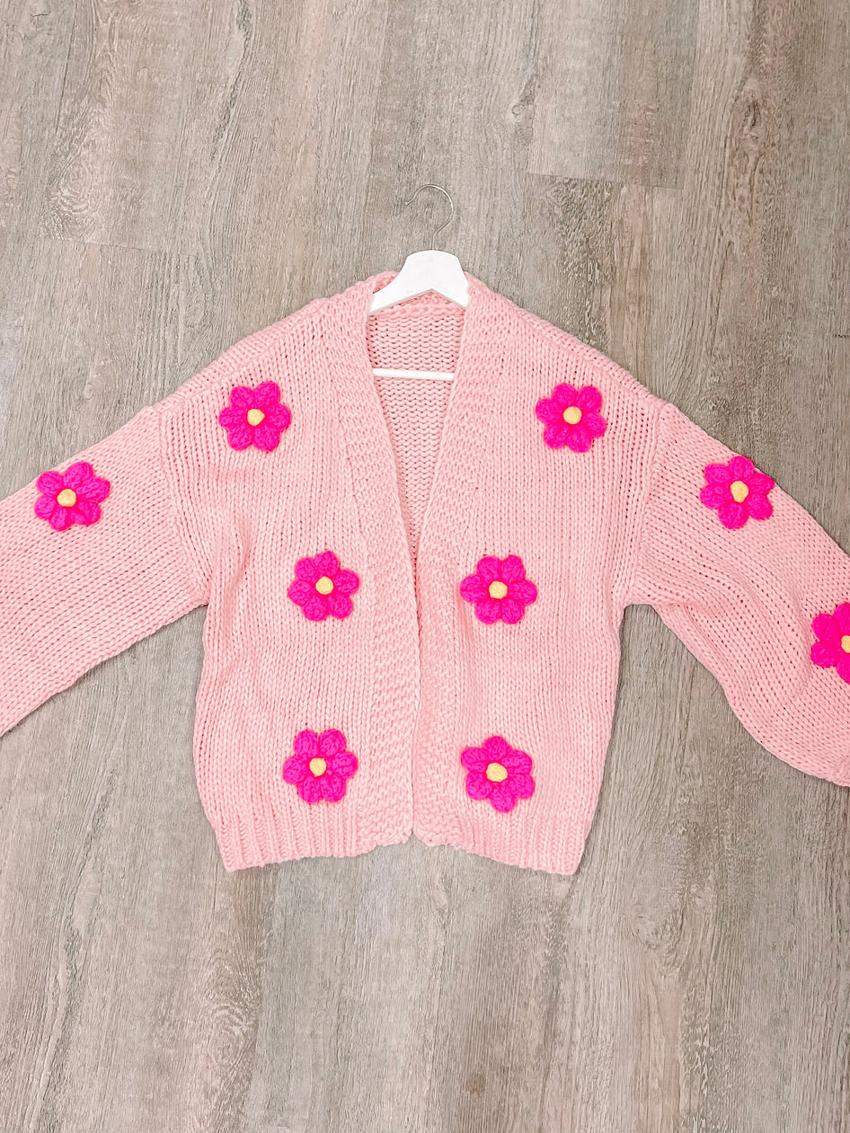 Pink knit cardigan adorned with bright pink flowers, hanging on a white hanger against a wood background.