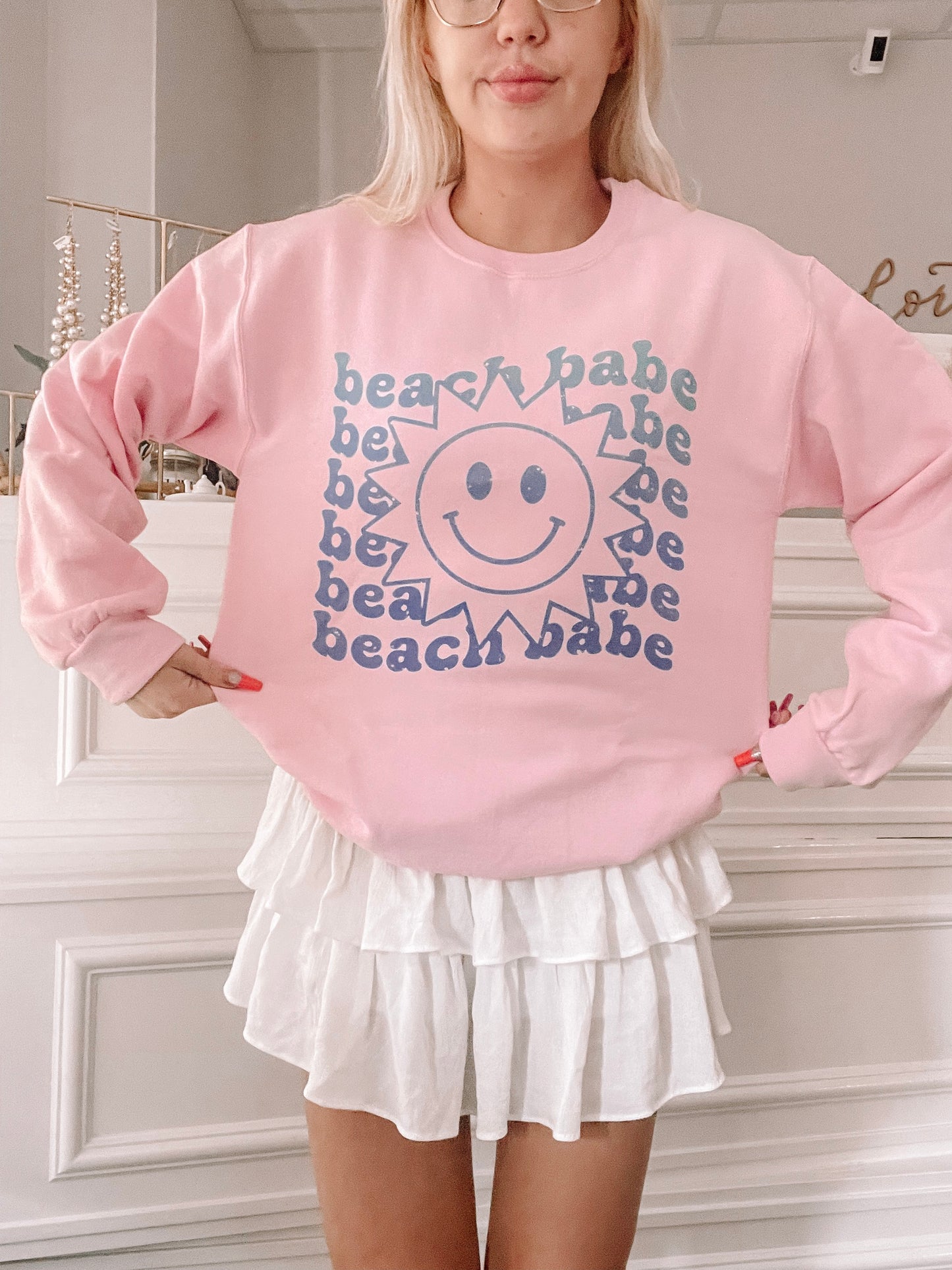 Pink sweatshirt featuring a smiling sun graphic and "beach babe" text, modeled indoors.