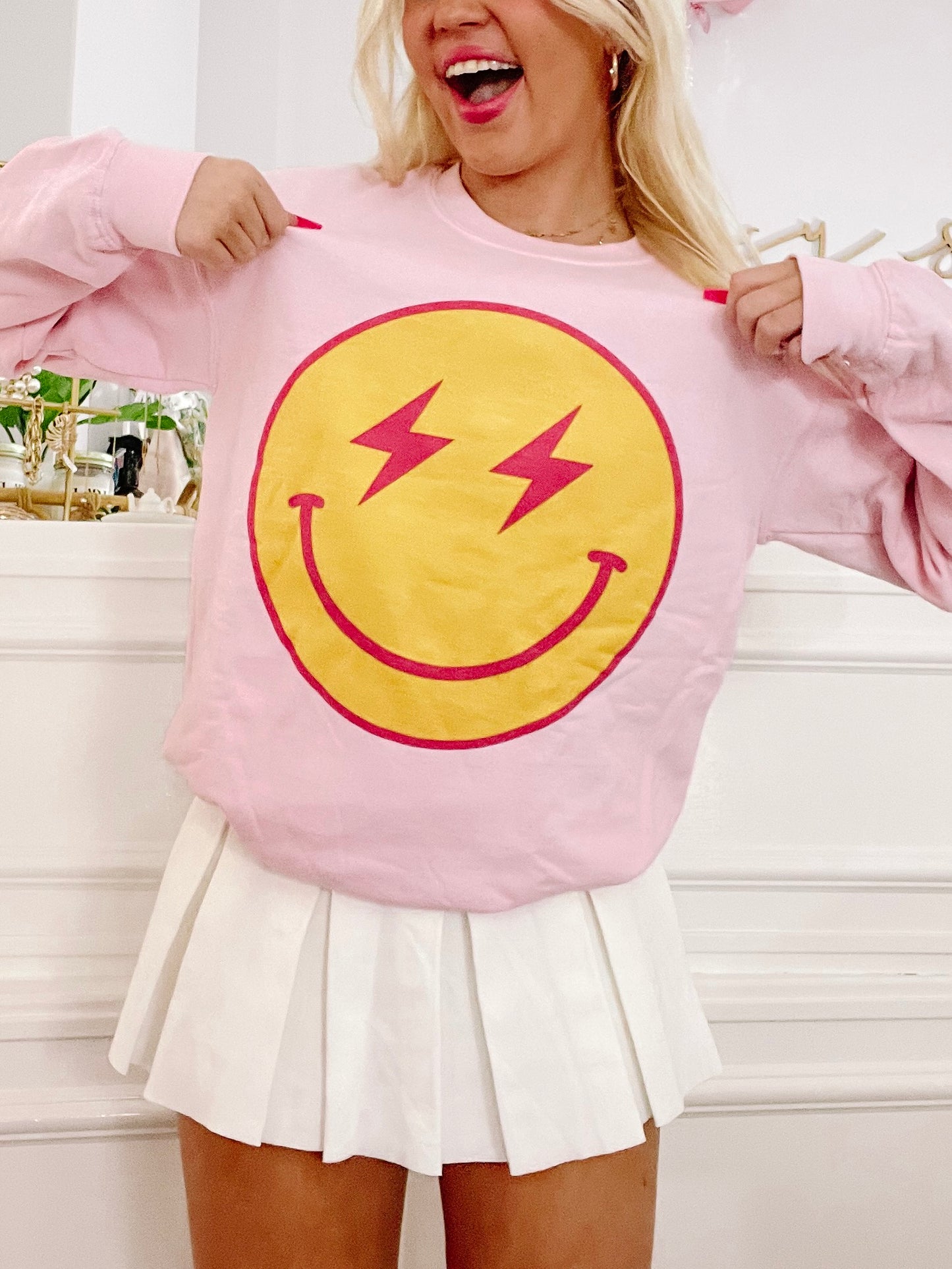 Pink crewneck sweatshirt featuring a vibrant yellow smiley face with red lightning bolts; model wearing with white pleated skirt.