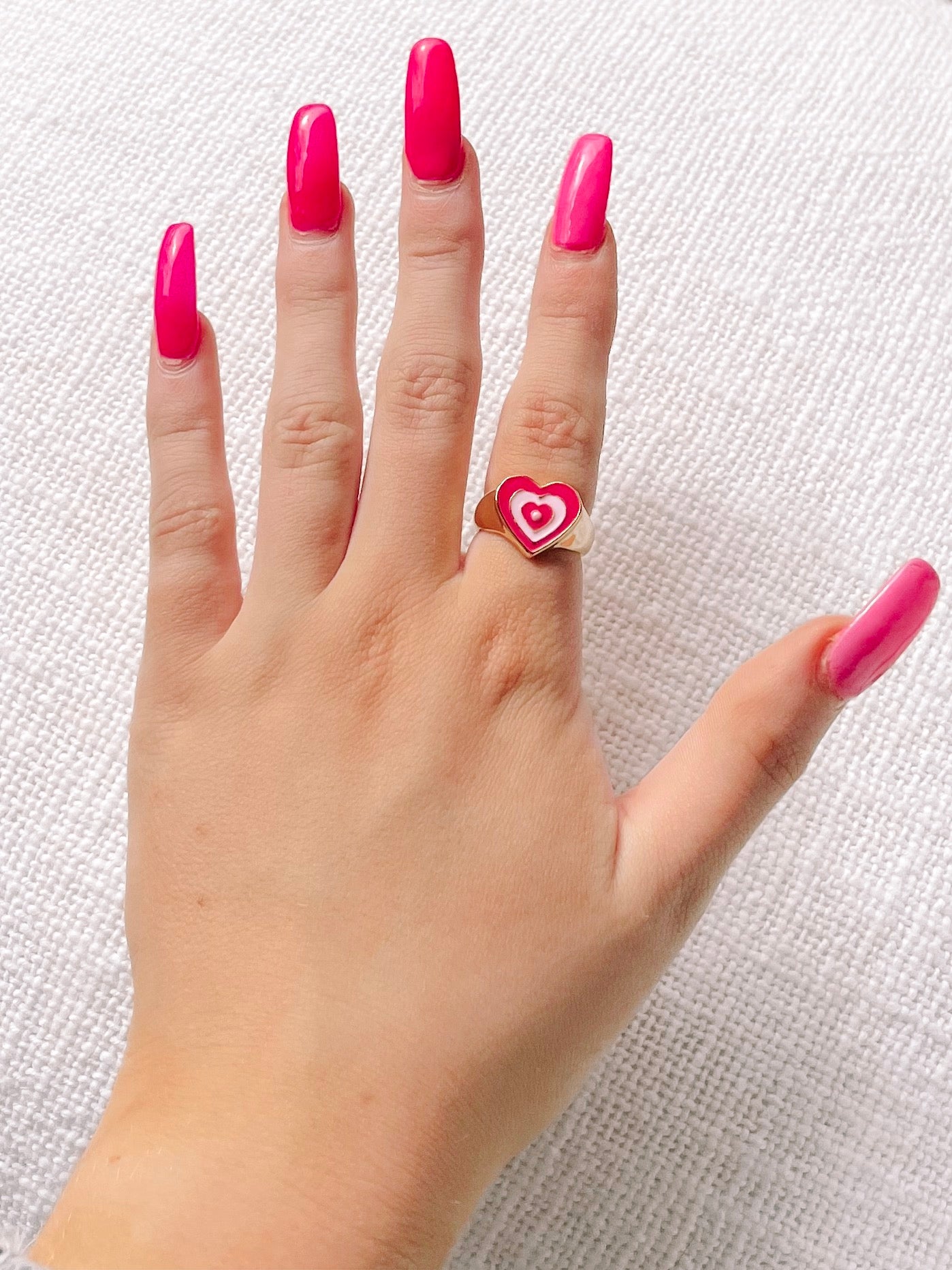 Gold ring with pink heart design, shown on a hand with bright pink nails.