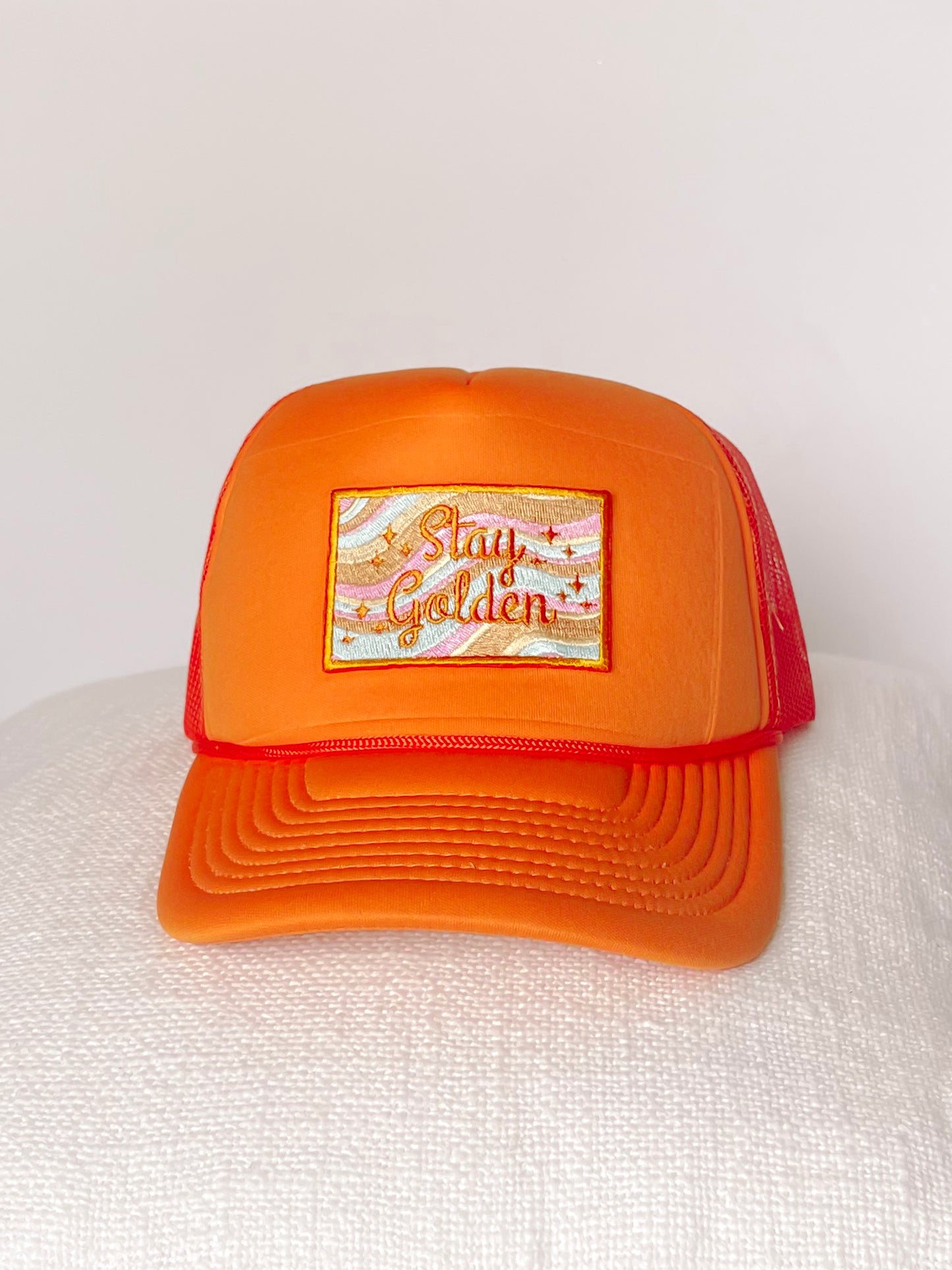 Orange trucker hat with embroidered "Stay Golden" patch; pastel wave design.